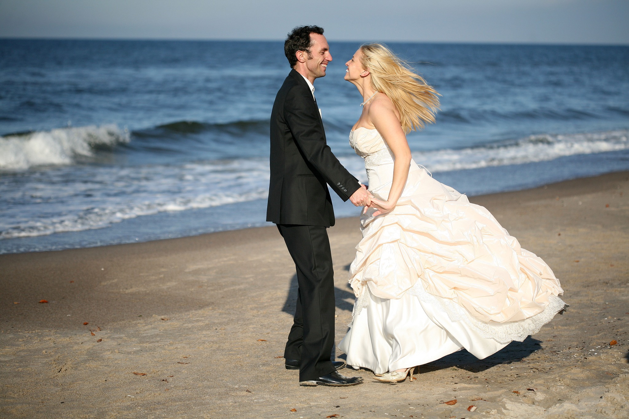 Uśmiechnięta para młoda na plaży, on w czarnym garniturze, ona w sukni ślubnej w odcieniach bieli i beżu, trzymają się za ręce na tle falującego morza.