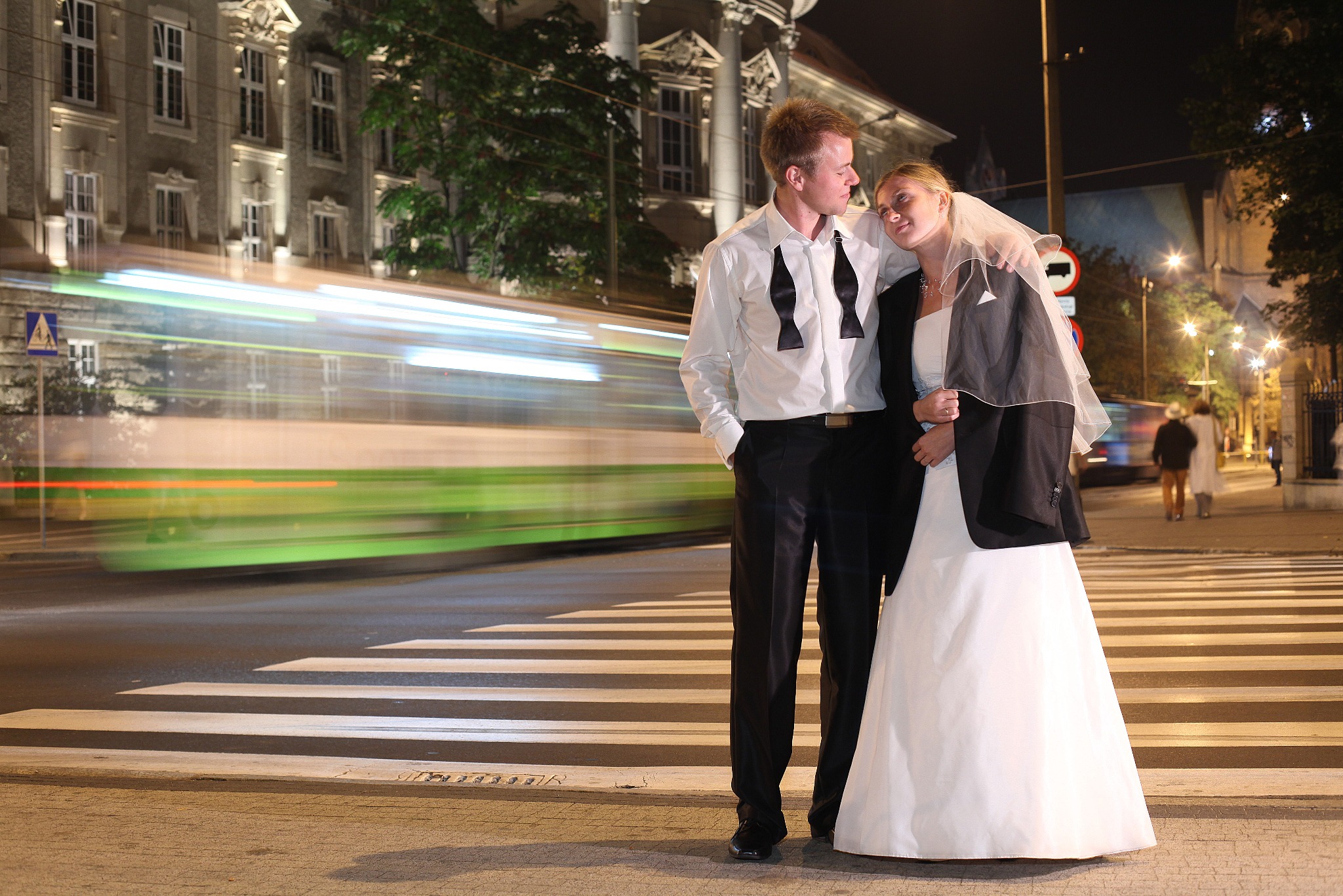 Para młoda na pasach nocą, z rozmytym tramwajem w tle. Pan młody w rozpiętej koszuli, panna młoda w sukni i marynarce. Romantyczna sesja w miejskiej scenerii.