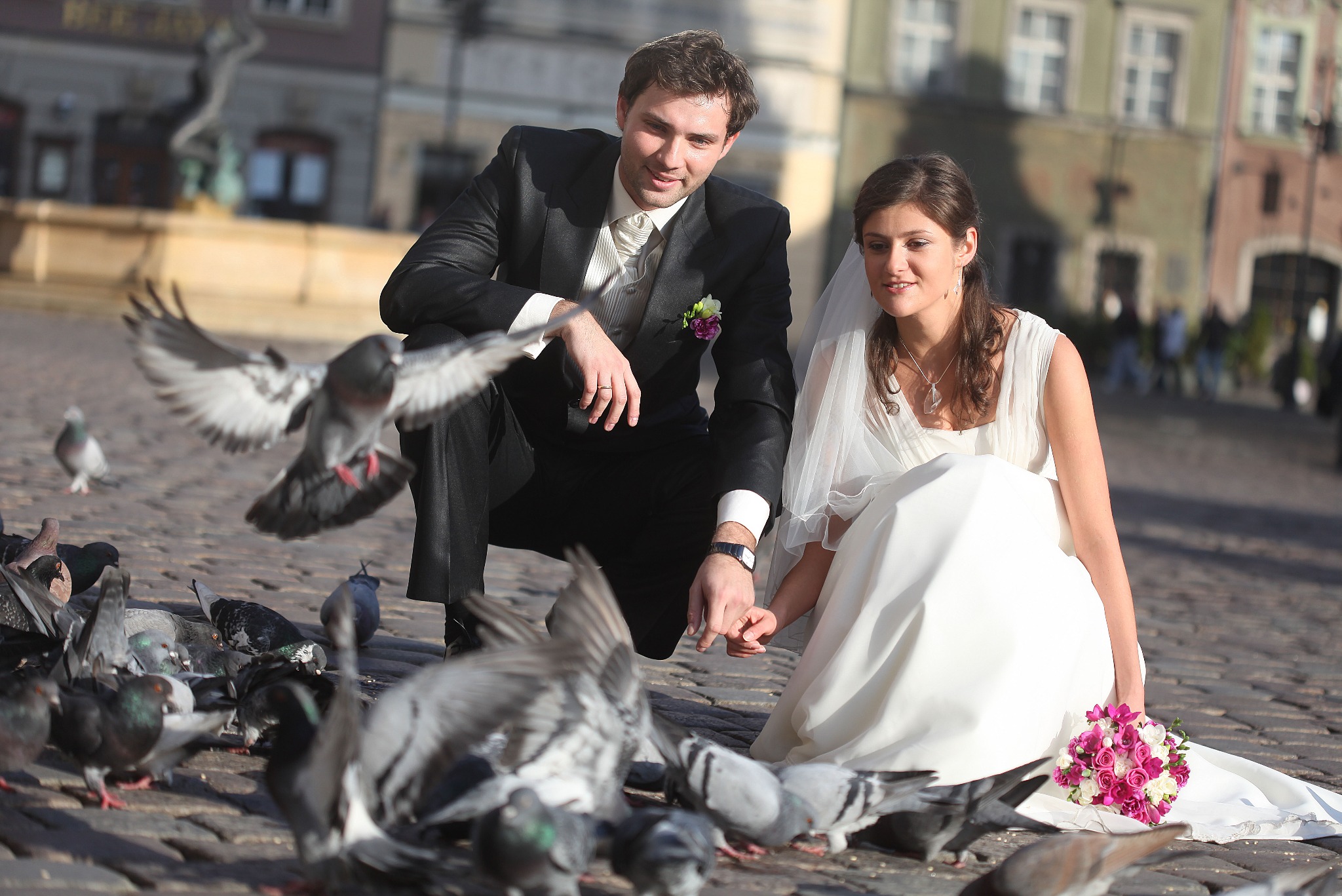 Para młoda w strojach ślubnych siedzi na brukowanym placu, otoczona przez stado gołębi w locie. W tle zabytkowe kamienice i fontanna.
