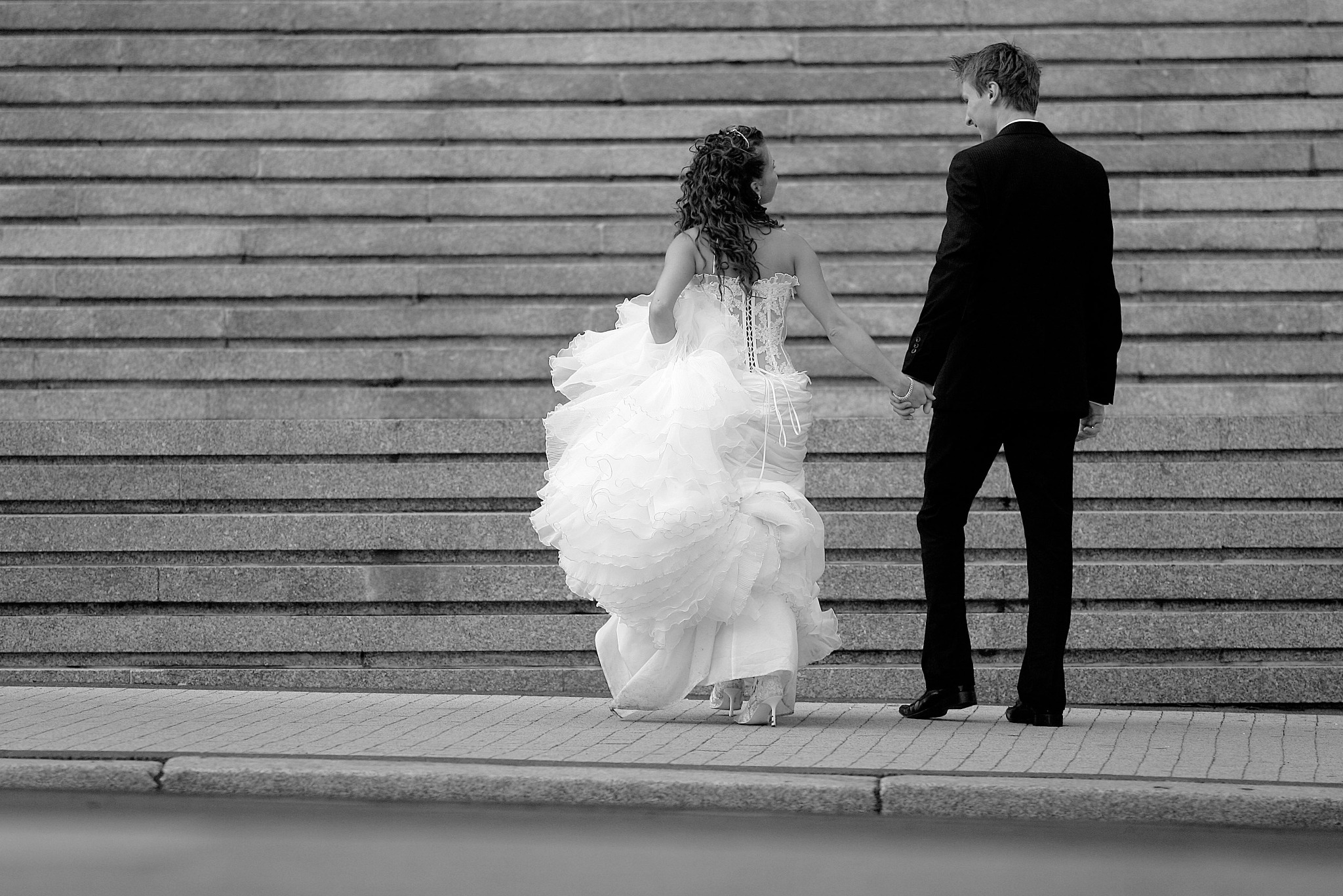 Czarno-białe zdjęcie pary młodej, idącej po schodach. Panna młoda w sukni z falbanami, pan młody w garniturze. Ujęcie z tyłu, skupione na detalach sukni i schodach.