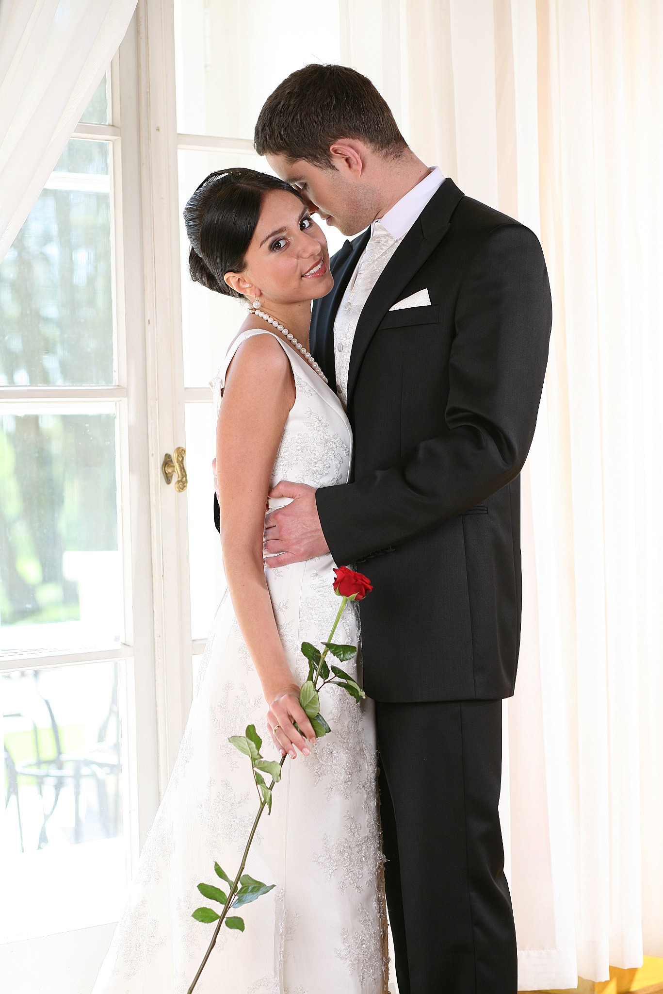 Uśmiechnięta panna młoda w białej sukni trzyma czerwoną różę, wtulona w pana młodego w czarnym garniturze na tle okna z białymi zasłonami.