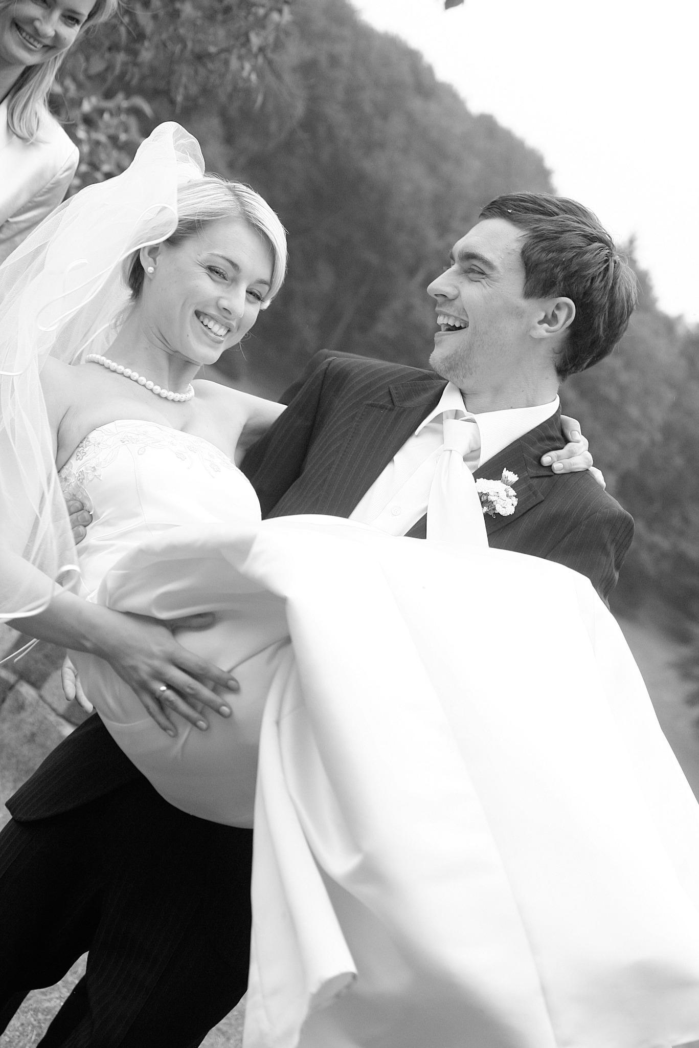 Czarno-białe zdjęcie: Uśmiechnięty pan młody niesie na rękach roześmianą pannę młodą w sukni ślubnej, w tle zieleń. Kadr z wesela, uchwycony moment radości.