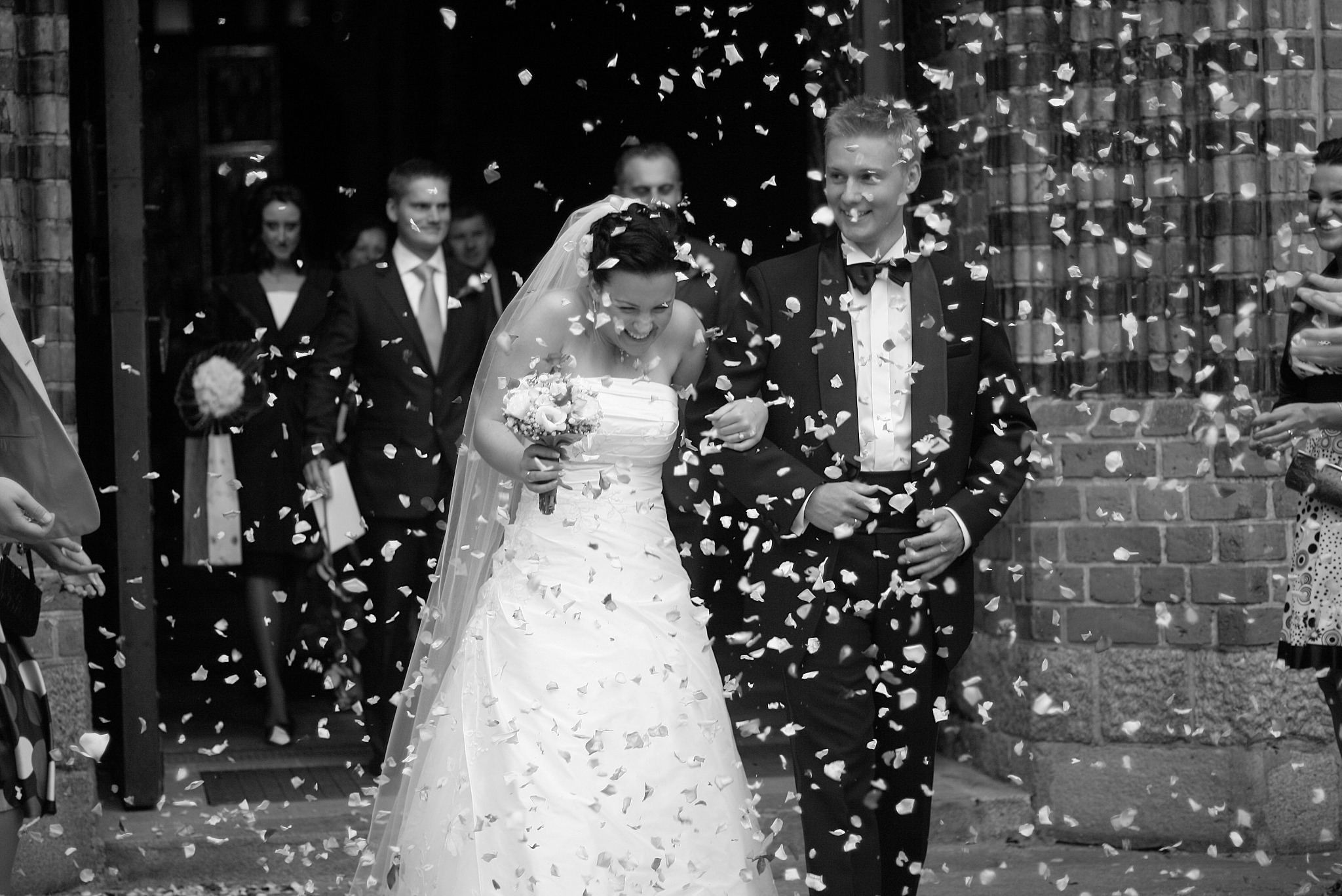 Czarno-białe zdjęcie uśmiechniętej pary młodej wychodzącej z kościoła, obsypywanej konfetti. W tle goście weselni i ceglana ściana.