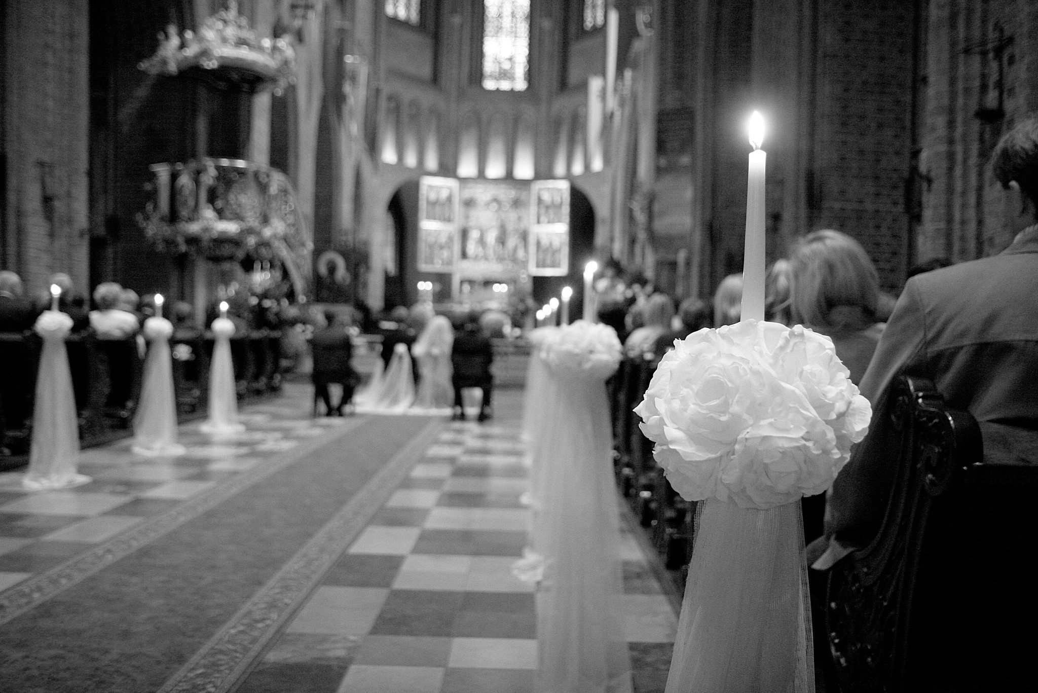 Czarno-białe zdjęcie wnętrza kościoła z dekoracjami kwiatowymi i świecami wzdłuż przejścia, gdzie w tle widać parę młodą podczas ceremonii ślubnej.
