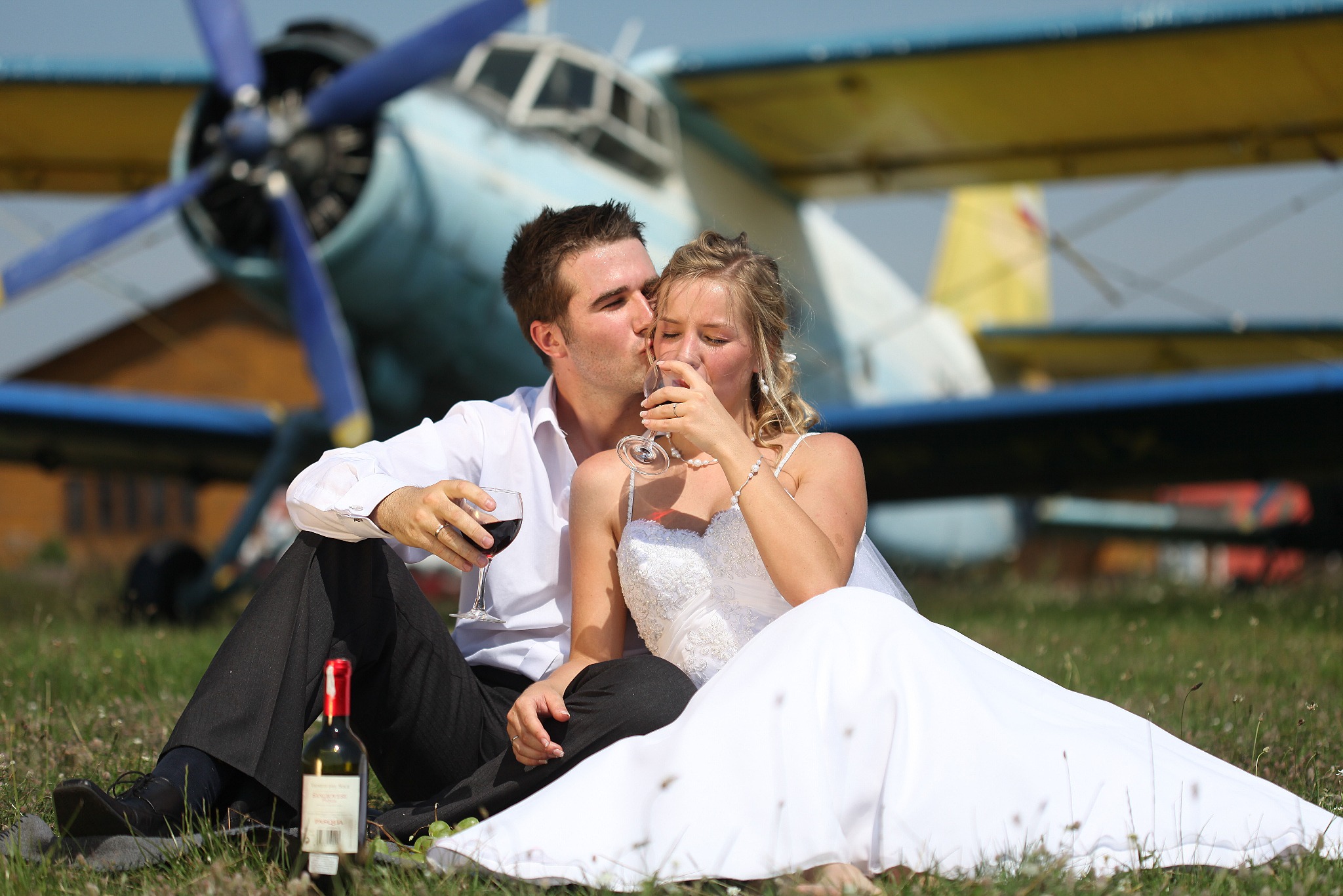 Para młoda siedzi na trawie z butelką wina i kieliszkami na tle samolotu. Mężczyzna całuje kobietę w policzek. Romantyczna sesja ślubna w plenerze.