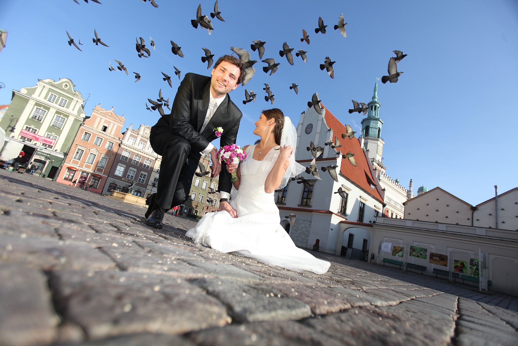Uśmiechnięta para młoda w centrum miasta, otoczona stadem gołębi. Pan młody w czarnym garniturze, panna młoda w białej sukni. Nietypowy kadr z niskiej perspektywy.