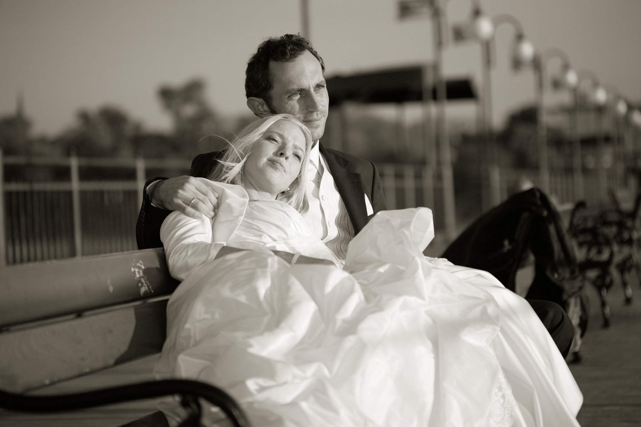 Sepia: Para młoda siedzi na ławce, on w garniturze, ona w sukni ślubnej. W tle latarnie i zieleń. Ujęcie z boku, z delikatnym światłem podkreślającym detale.