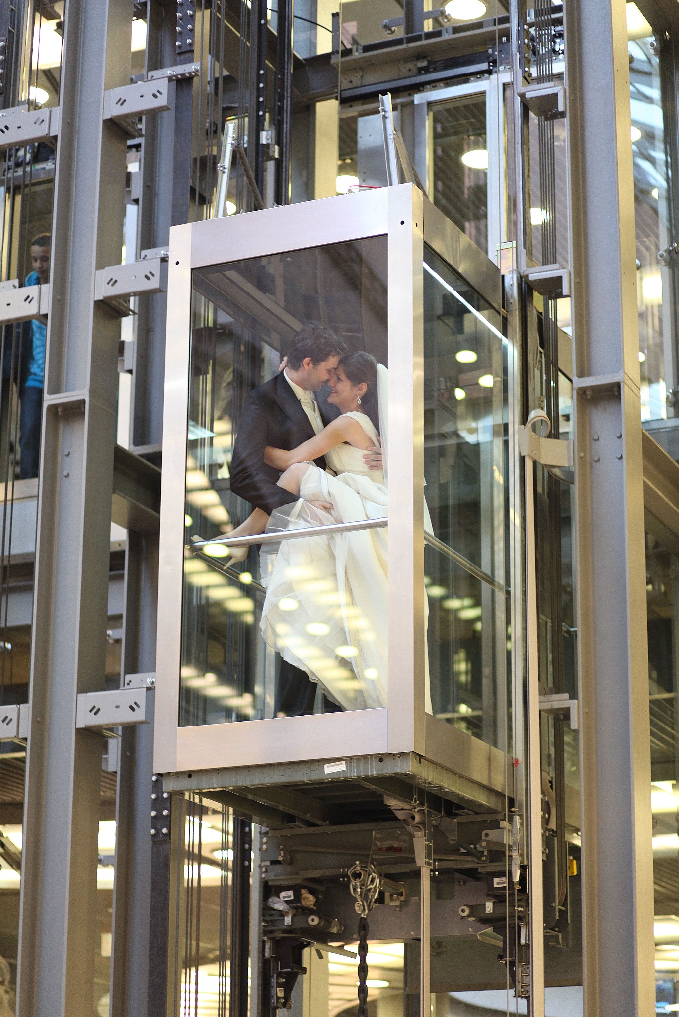 Para młoda w objęciach w szklanej windzie na tle metalowej konstrukcji. Ujęcie z dołu, podkreślające nowoczesny charakter sesji ślubnej.