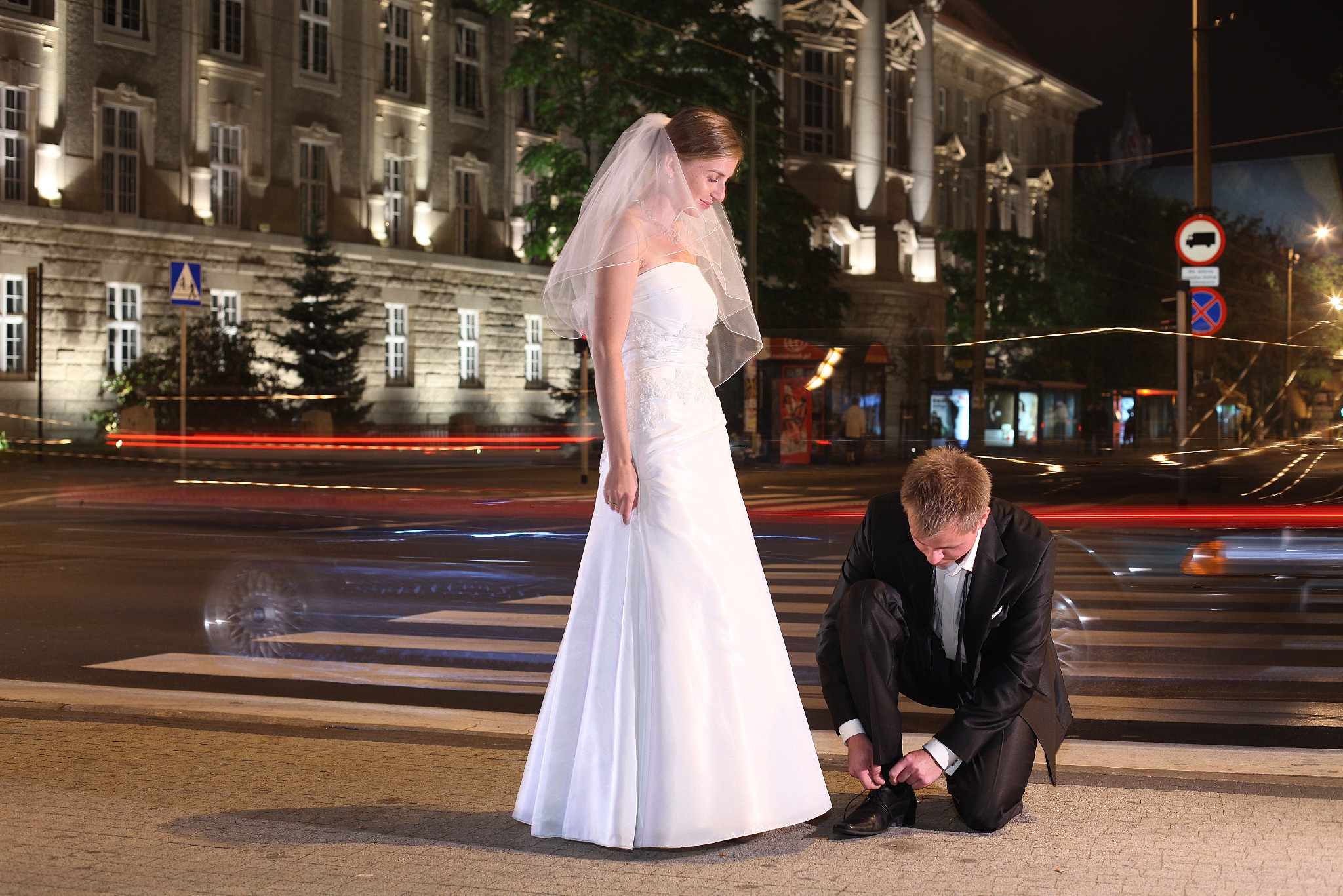 Panna młoda w białej sukni i welonie stoi na pasach, a pan młody klęczy obok, zawiązując sznurówki. W tle nocne miasto z rozmytymi światłami samochodów.