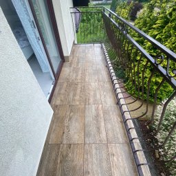 ST MASTER Anatolii Pavlenko i Oleksandr Sliuzka spółka cywilna - Balkon z brązowymi płytkami imitującymi drewno, metalowa balustrada. Widok na zieleń. Płytki ułożone w regularny wzór, fuga w kolorze zbliżonym do płytek.