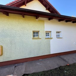 ST MASTER Anatolii Pavlenko i Oleksandr Sliuzka spółka cywilna - Malowanie elewacji domu: fragment ściany w dwóch kolorach (żółty i biały), malarz z wałkiem w trakcie pracy. Widoczne zabezpieczone okna i skrzynka gazowa.