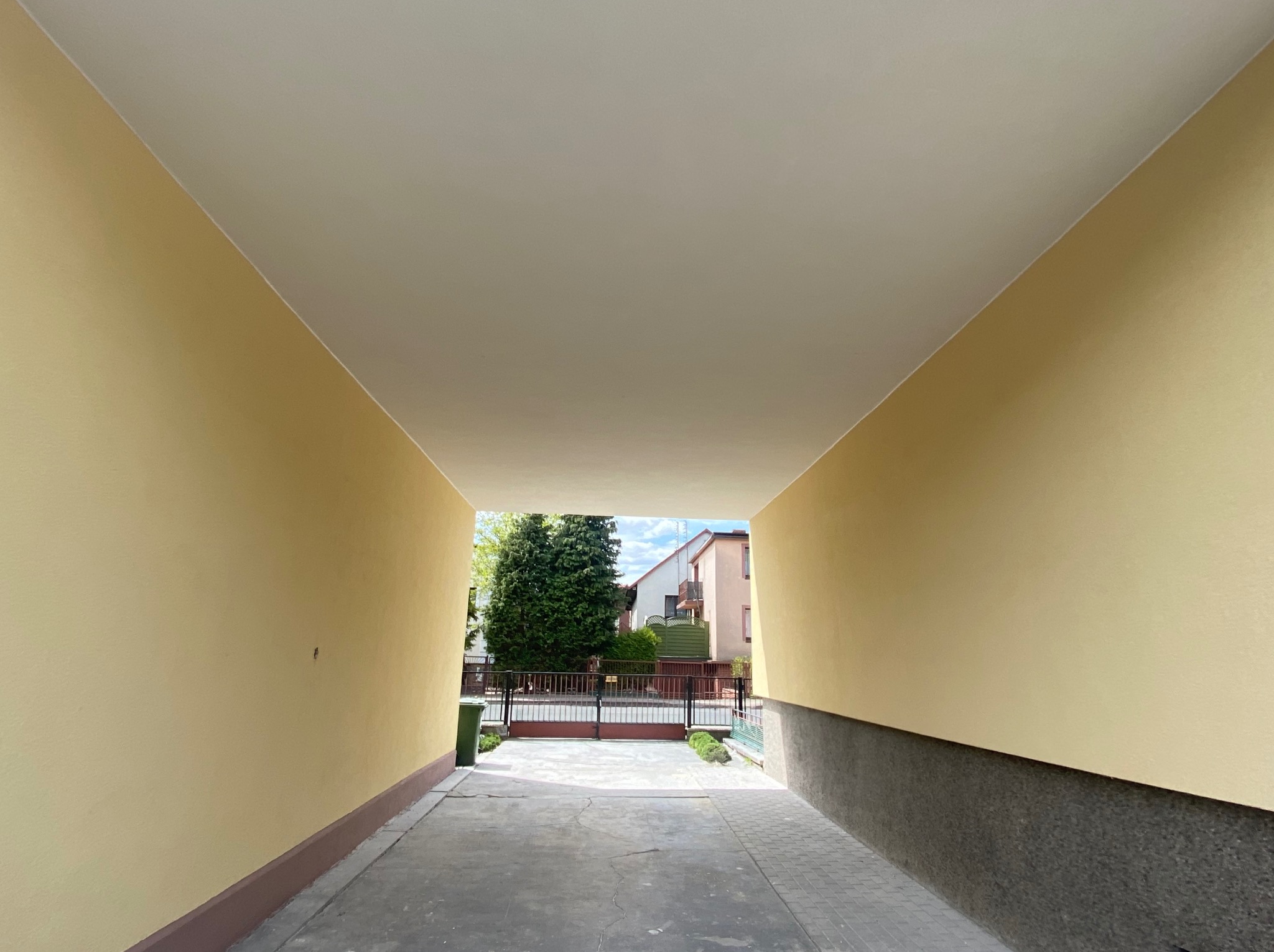 Pomalowane na jasnożółto ściany w przejściu do bramy wjazdowej. Widoczne świeże malowanie, szara podmurówka i brama w tle.