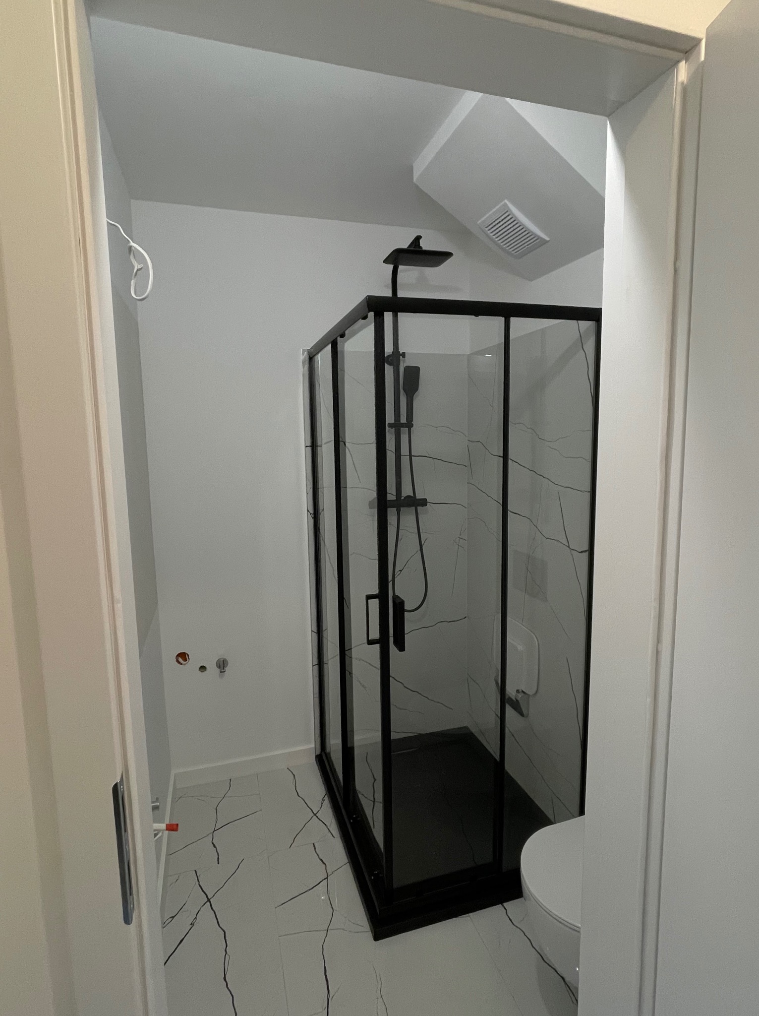 Nowoczesna kabina prysznicowa z czarnymi ramami i marmurowym wzorem na szkle, widoczna zza uchylonych drzwi łazienki. Białe płytki podłogowe z czarnymi żyłkami.