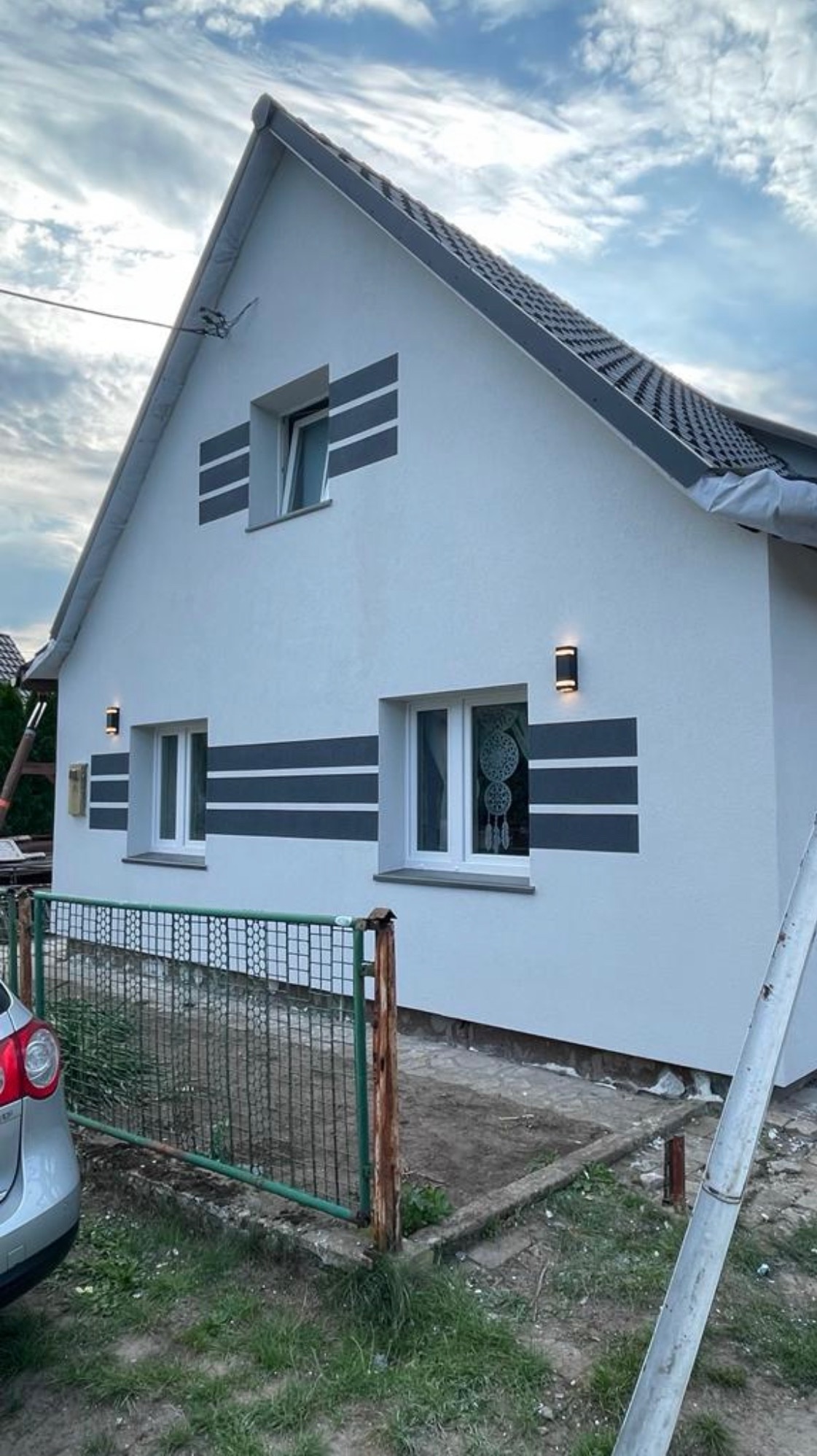 Elewacja domu jednorodzinnego po malowaniu w Szczecinie. Biała fasada z szarymi pasami, oświetlenie zewnętrzne. Widok z zewnątrz, ogrodzenie i fragment samochodu.