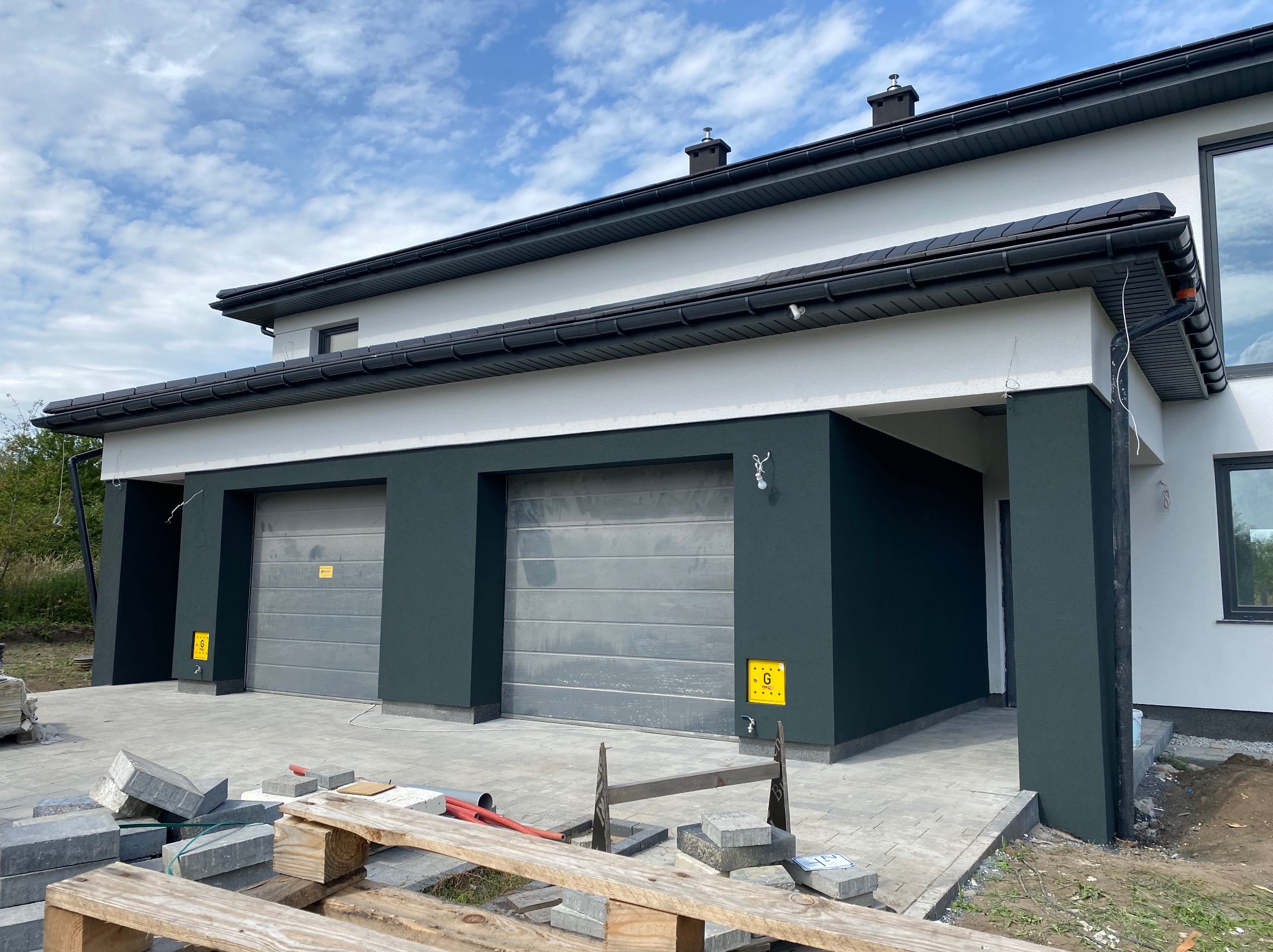 Nowoczesny dom jednorodzinny z dwoma garażami, wykończony w bieli i ciemnej zieleni. Na pierwszym planie kostka brukowa i materiały budowlane.