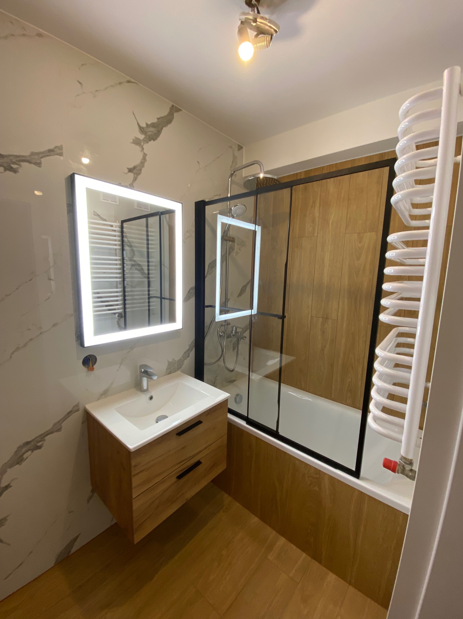 Nowoczesna łazienka z drewnianymi akcentami: szafka pod umywalką, panele prysznicowe. Lustro z podświetleniem LED, grzejnik drabinkowy.