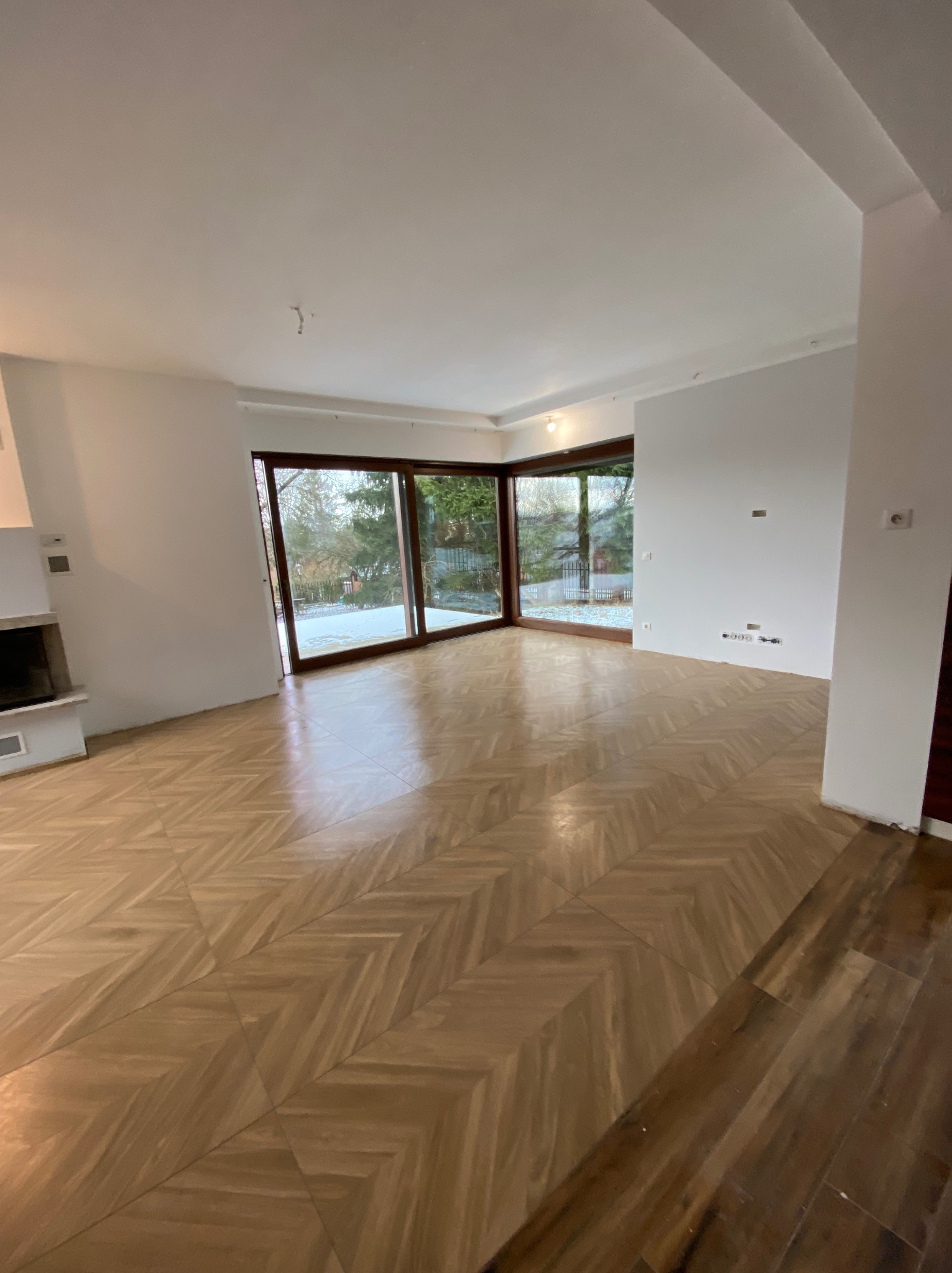 Jasny parkiet ułożony we wzór jodełki francuskiej w przestronnym, odnowionym salonie z dużymi oknami i kominkiem. Świeżo pomalowane ściany, widok na zimowy ogród.