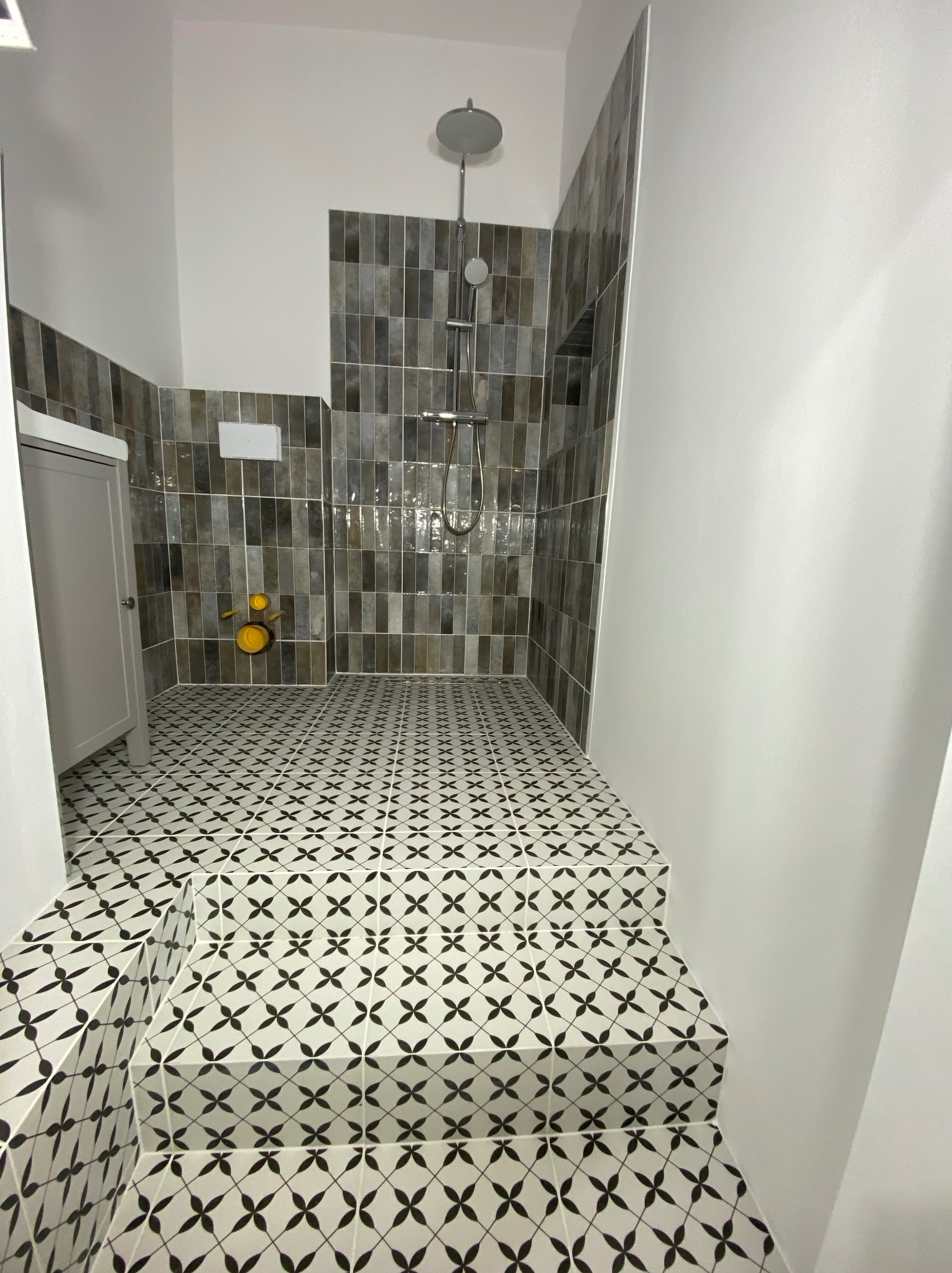 Nowoczesna łazienka z kabiną prysznicową typu walk-in, wykończona szaro-brązowymi płytkami na ścianach i wzorzystą, czarno-białą mozaiką na podłodze i schodach.