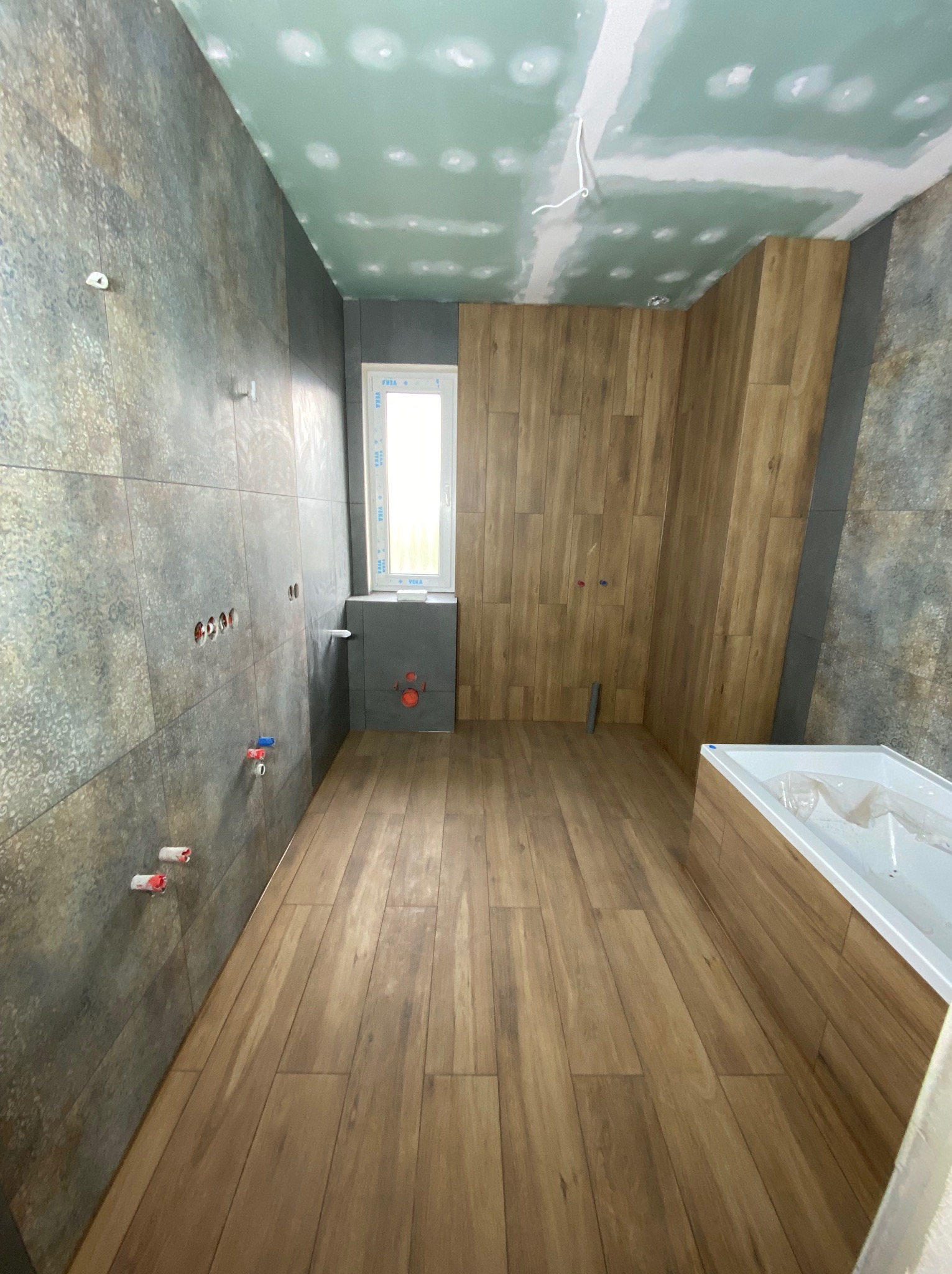 Wnętrze łazienki w trakcie remontu: położone płytki imitujące drewno na podłodze i ścianie, sufit z płyt gipsowo-kartonowych, przygotowane instalacje wod-kan.