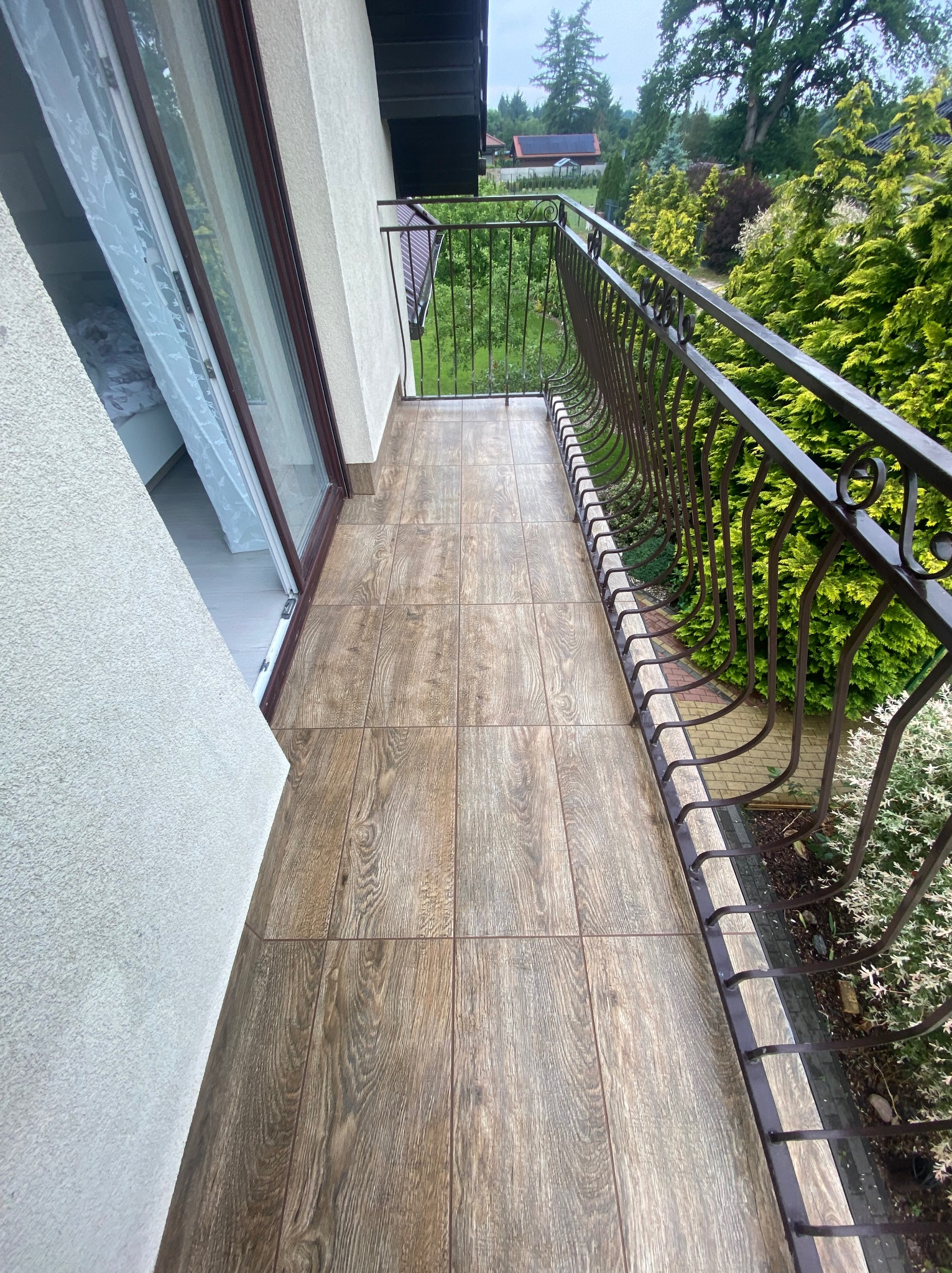 Balkon z brązowymi płytkami imitującymi drewno, metalowa balustrada. Widok na zieleń. Płytki ułożone w regularny wzór, fuga w kolorze zbliżonym do płytek.