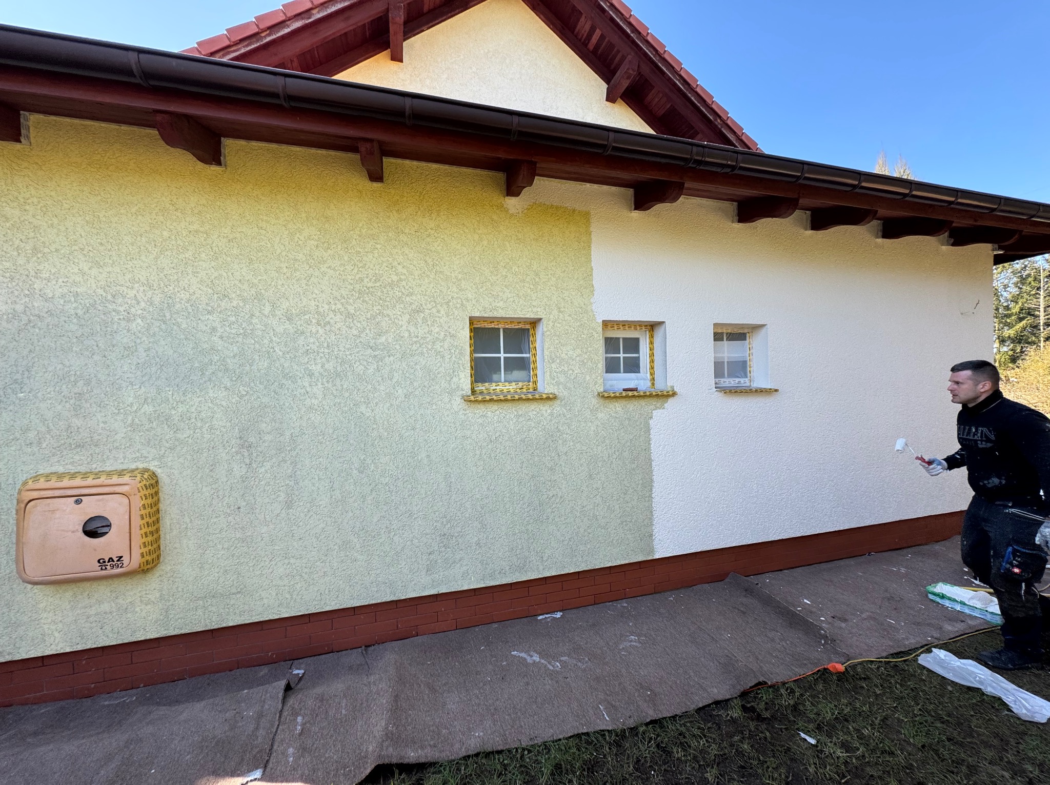 Malowanie elewacji domu: fragment ściany w dwóch kolorach (żółty i biały), malarz z wałkiem w trakcie pracy. Widoczne zabezpieczone okna i skrzynka gazowa.