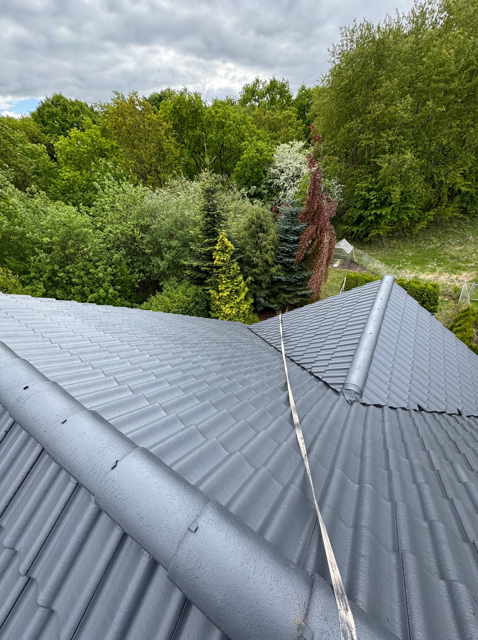 Szary dach z dachówki ceramicznej, widok z góry na tle zieleni drzew i pochmurnego nieba. Widoczny komin i taśma zabezpieczająca.