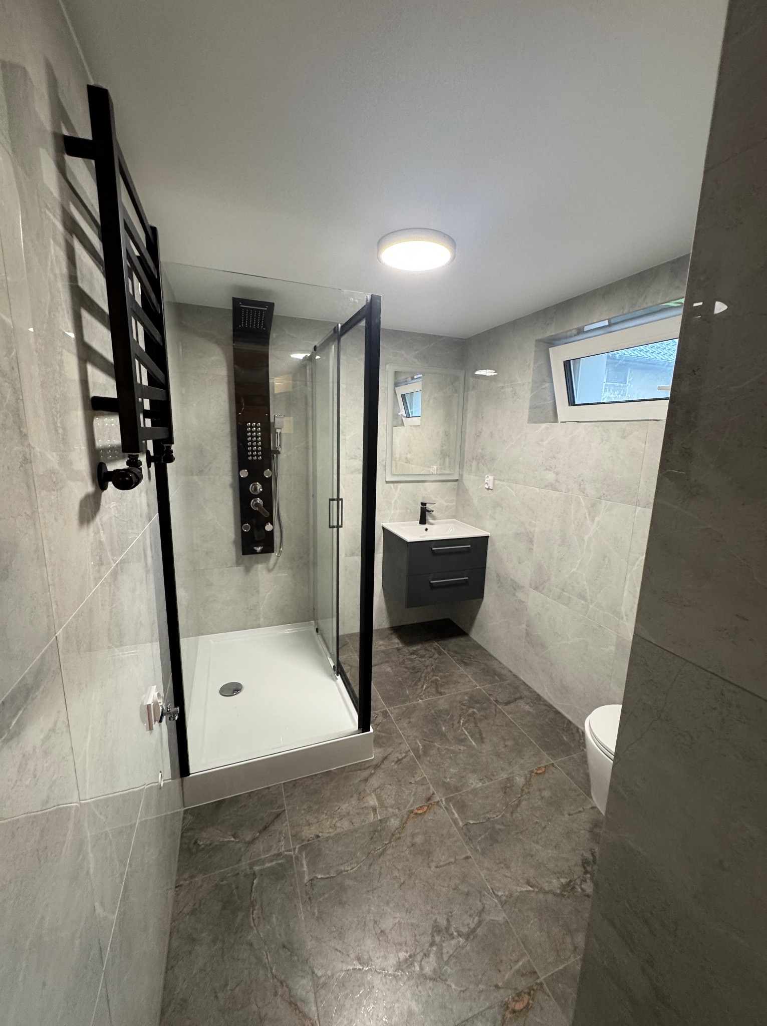 Nowoczesna łazienka z kabiną prysznicową typu walk-in, szarymi płytkami imitującymi marmur, czarną armaturą i podwieszaną szafką z umywalką. Minimalistyczny design.