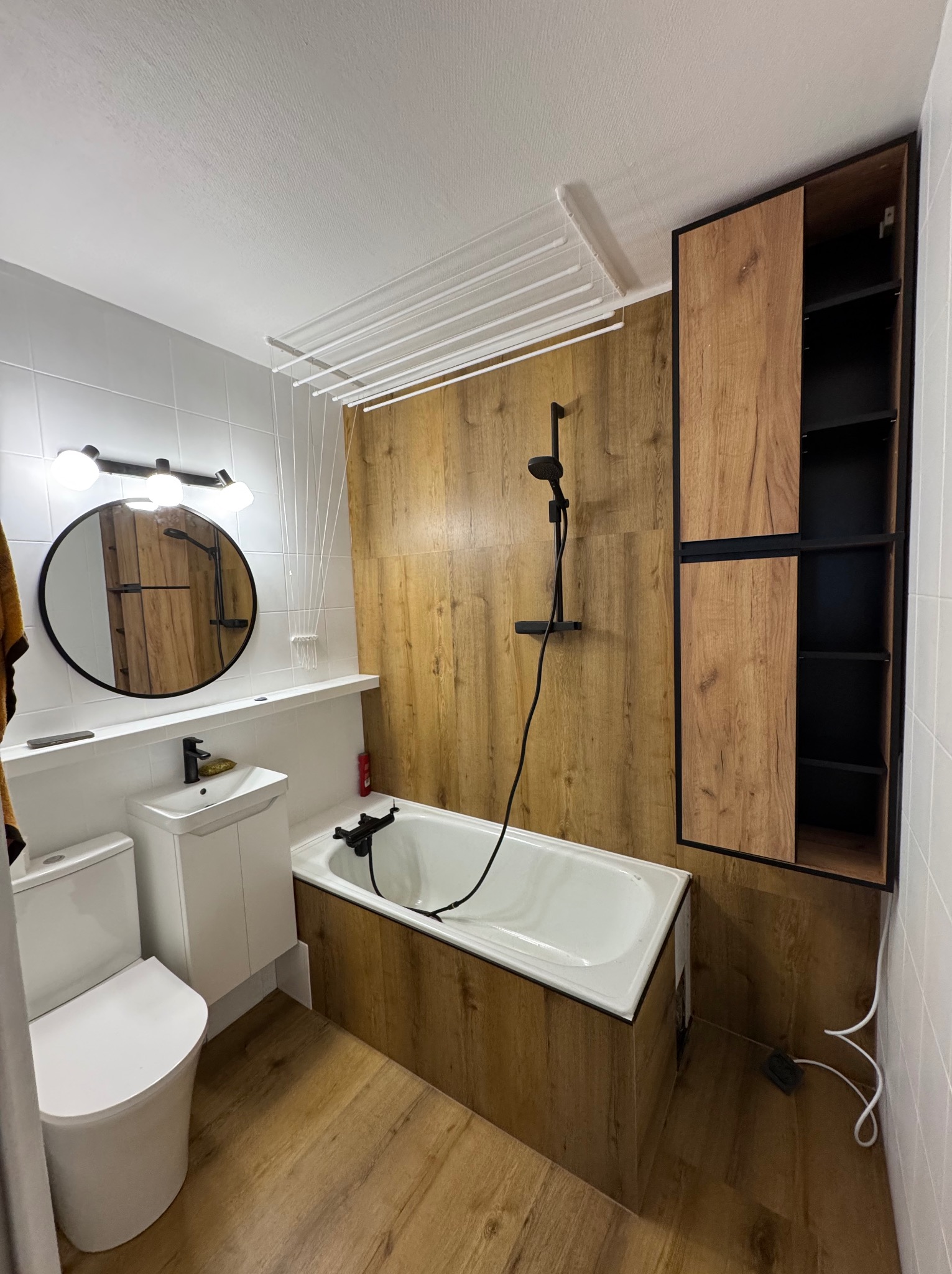 Nowoczesna łazienka z drewnopodobnymi płytkami, białą armaturą i czarnymi akcentami. Widoczna wanna, umywalka z szafką, toaleta i szafka wisząca.