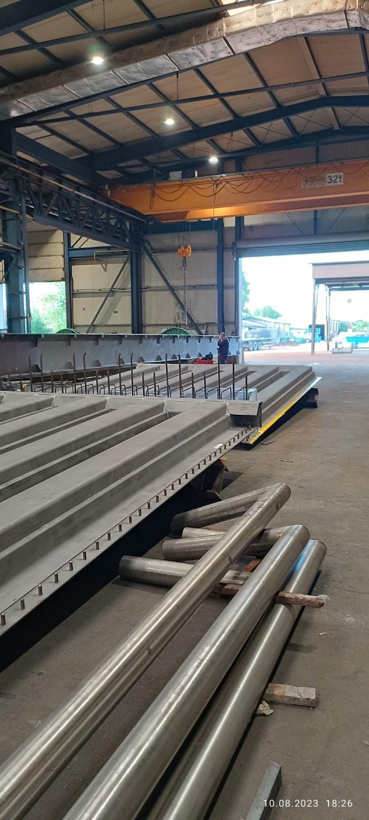 Prefabrykowane elementy betonowe z wystającymi prętami zbrojeniowymi w hali produkcyjnej, widoczne rury i suwnica. Data i godzina w prawym dolnym rogu.