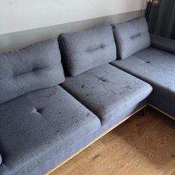 NCLEAN - mobilne pranie tapicerki - Szara, narożna kanapa z widocznymi plamami na siedzisku, ukazana z perspektywy pod kątem. Drewniana podłoga i fragment lampy w tle.