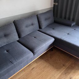 NCLEAN - mobilne pranie tapicerki - Szara, modułowa kanapa z drewnianym wykończeniem, ustawiona na parkiecie. W tle szare zasłony i lampa podłogowa. Na ścianie wiszą obrazy w czarnych ramkach.