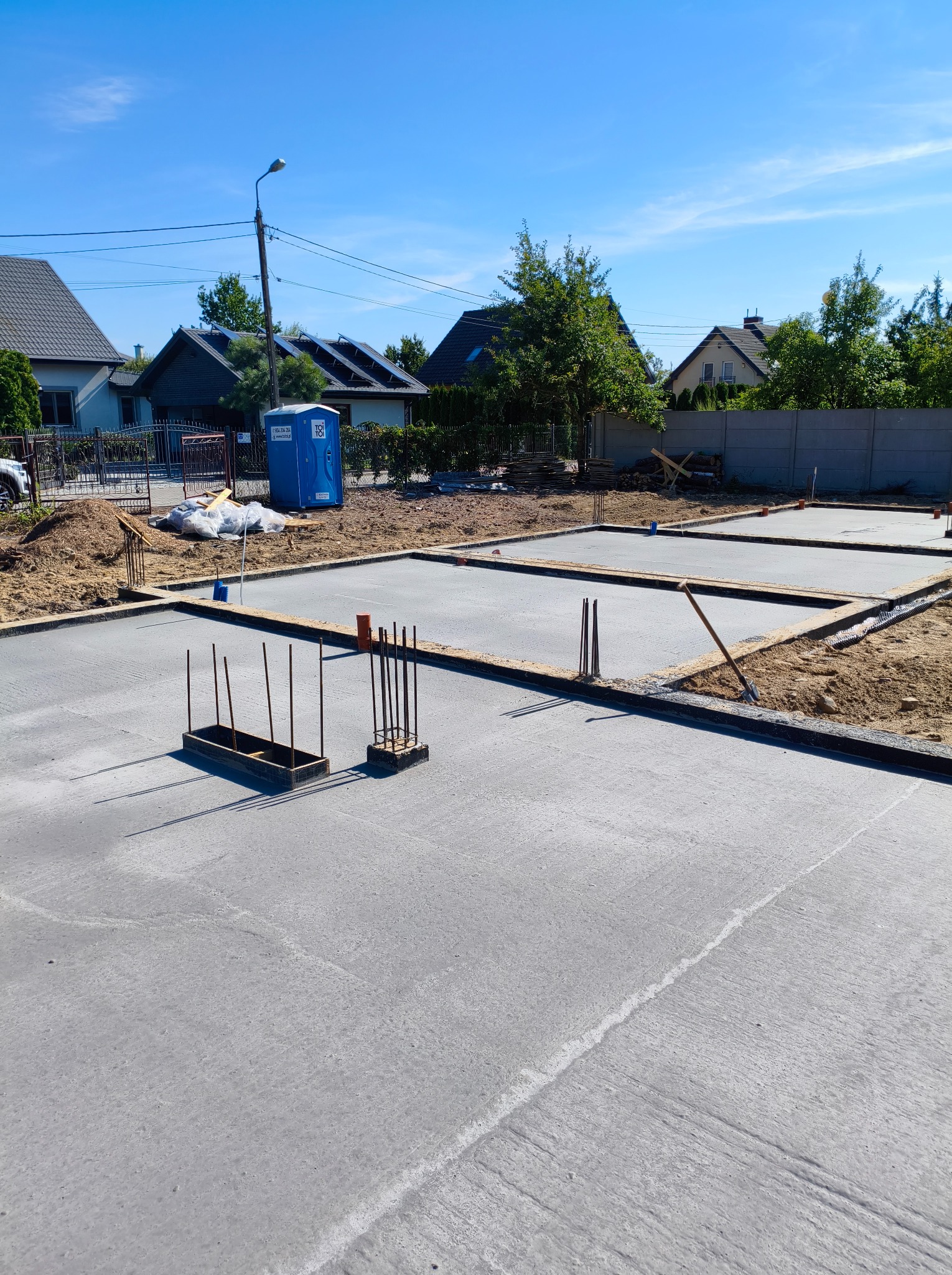 Wylana płyta fundamentowa z wystającymi prętami zbrojeniowymi na budowie domu jednorodzinnego w słoneczny dzień. Widoczne szalunki i TOI TOI.