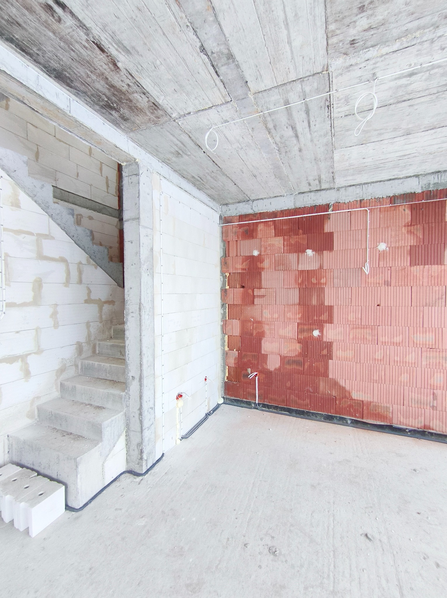 Wnętrze w stanie surowym: ściany z czerwonej cegły i białego betonu komórkowego, betonowe schody, widoczne instalacje elektryczne. Surowy betonowy strop.