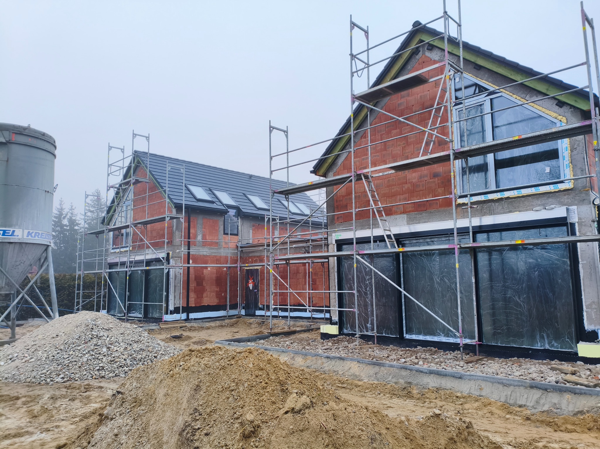 Dwa domy w trakcie budowy, z ceglanymi ścianami i rusztowaniami. Widoczne okna zabezpieczone folią, hałdy piasku i żwiru. Pochmurne niebo w tle.