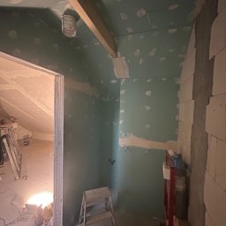 Igor Rędziak - Poddasze w trakcie adaptacji: ściany z zielonej płyty gipsowej, widoczne łączenia i szpachlowanie. Fragment konstrukcji drewnianej i drabina w pomieszczeniu.