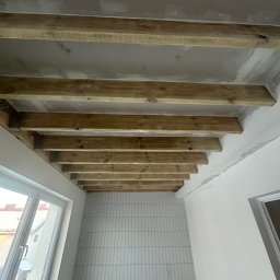 Igor Rędziak - Drewniana konstrukcja sufitu podwieszanego w trakcie montażu, widok z dołu. Ściany przygotowane pod wykończenie, widoczne puszki elektryczne i okno.