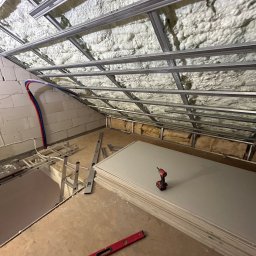 Igor Rędziak - Montaż sufitu podwieszanego z płyt gipsowo-kartonowych na poddaszu. Widoczne profile, izolacja, płyty i narzędzia. Ujęcie z dołu, ukośne.