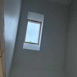 Igor Rędziak - Ściana poddasza po malowaniu na szaro, z oknem dachowym w drewnianej ramie. Widoczny fragment drewnianej konstrukcji. Gładka powierzchnia ściany.