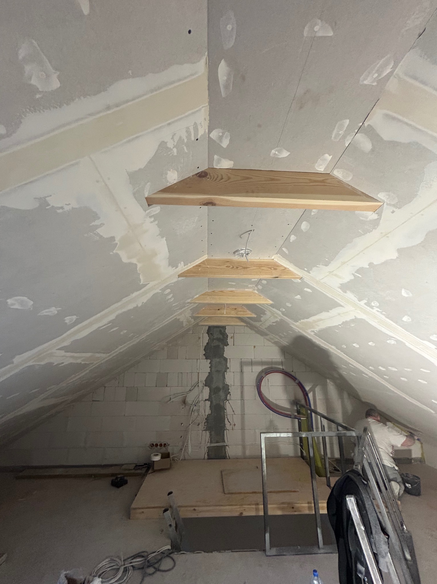 Poddasze w trakcie wykańczania: płyty karton-gips na skosach, drewniane elementy dekoracyjne pod sufitem, widoczne instalacje i narzędzia. Prace wykończeniowe w toku.