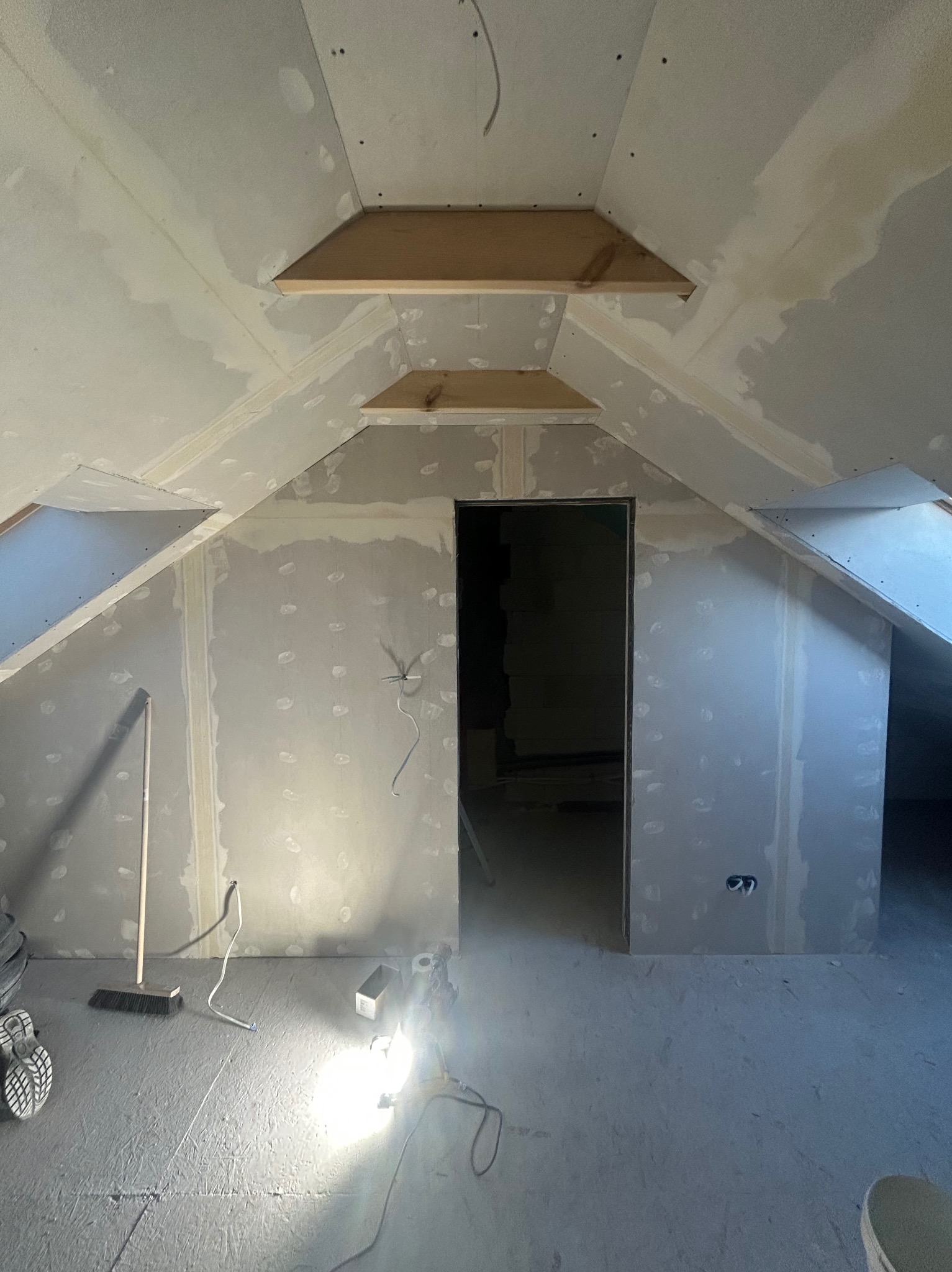 Poddasze w trakcie prac wykończeniowych: szpachlowane ściany z płyt karton-gips, drewniane elementy konstrukcyjne, widoczne narzędzia i oświetlenie budowlane.