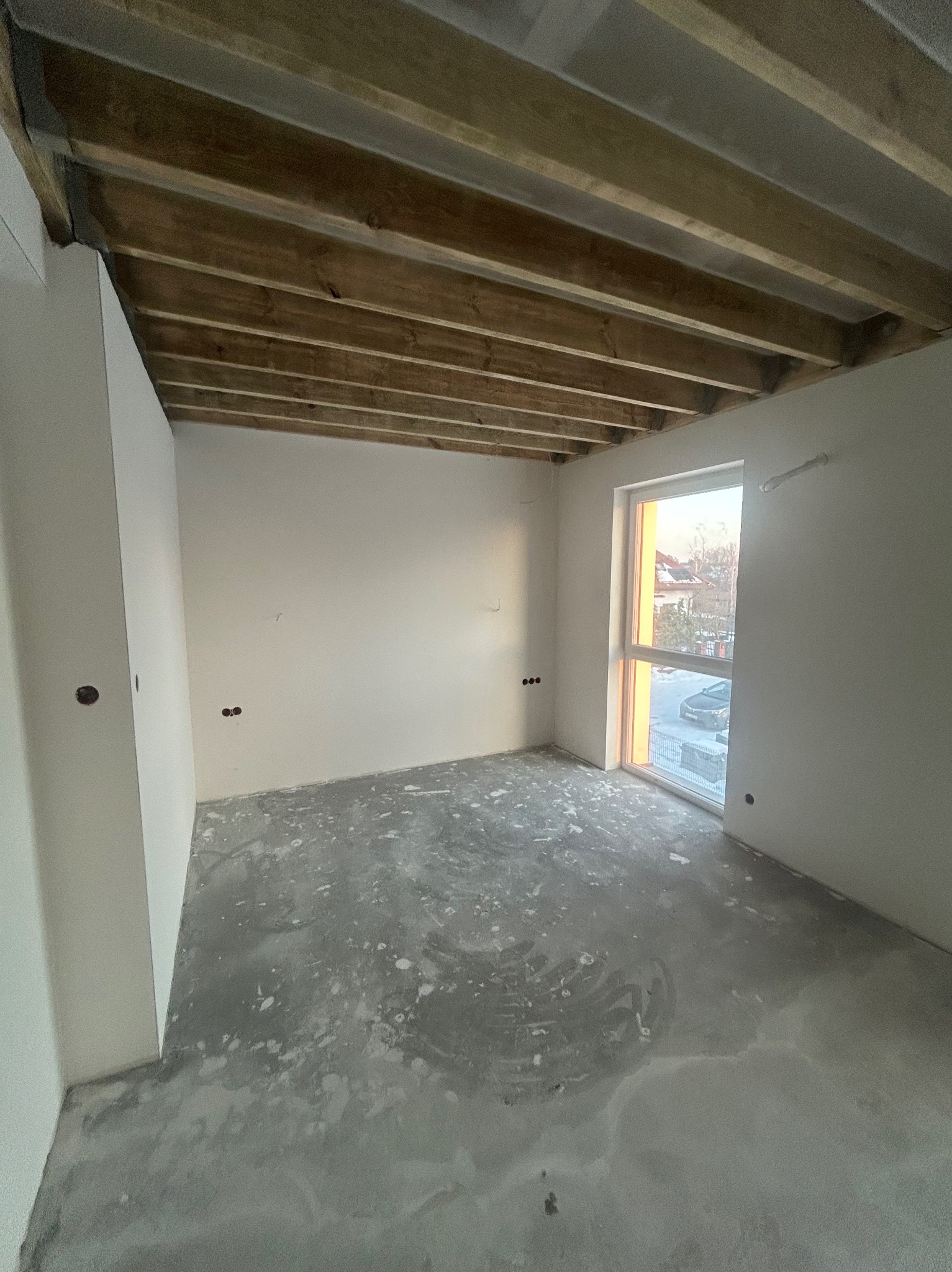 Surowe wnętrze pokoju z drewnianym stropem, gładkimi ścianami i betonową podłogą w trakcie wykańczania. Widoczne puszki elektryczne i okno z widokiem na zewnątrz.