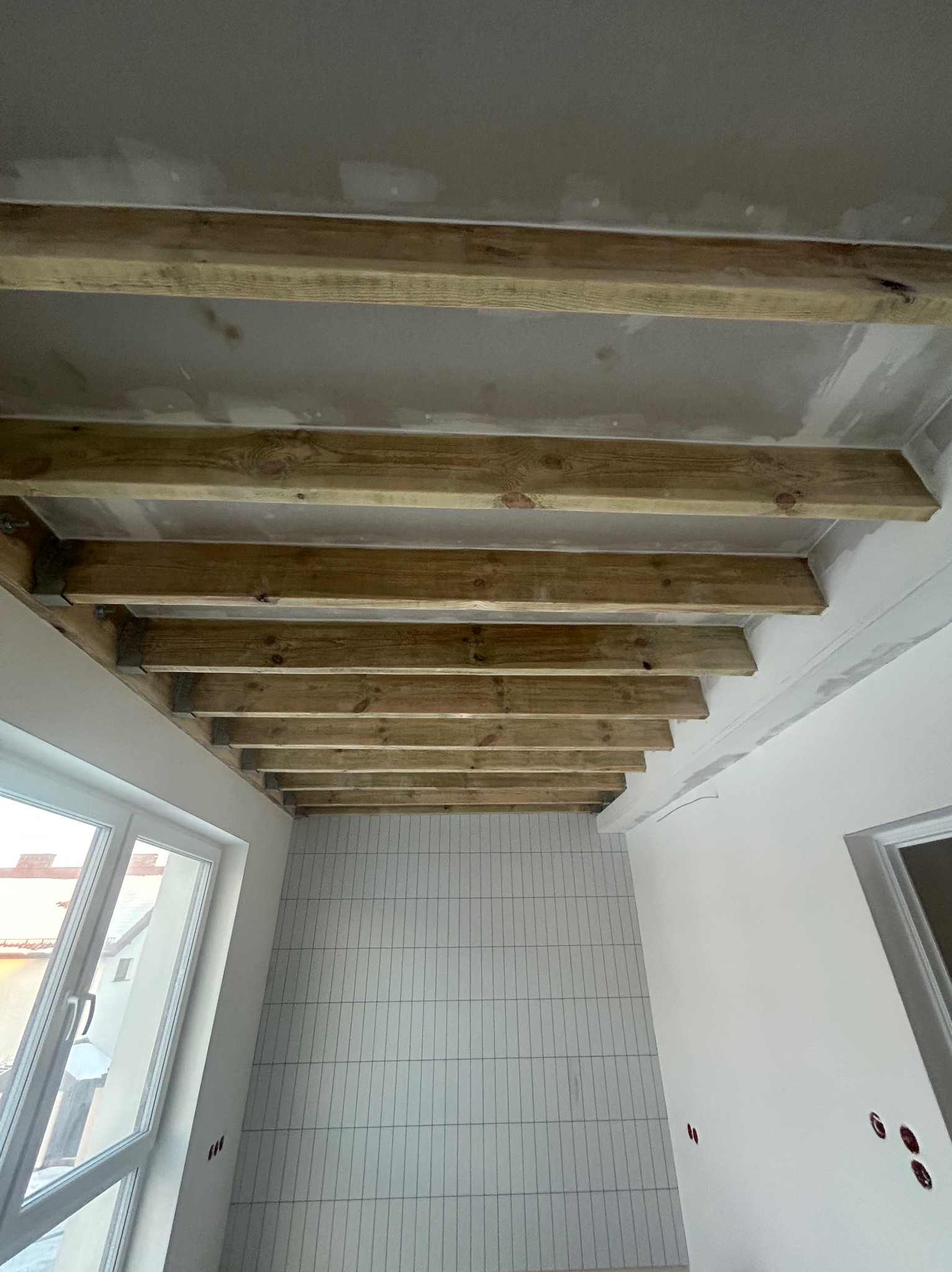 Drewniana konstrukcja sufitu podwieszanego w trakcie montażu, widok z dołu. Ściany przygotowane pod wykończenie, widoczne puszki elektryczne i okno.