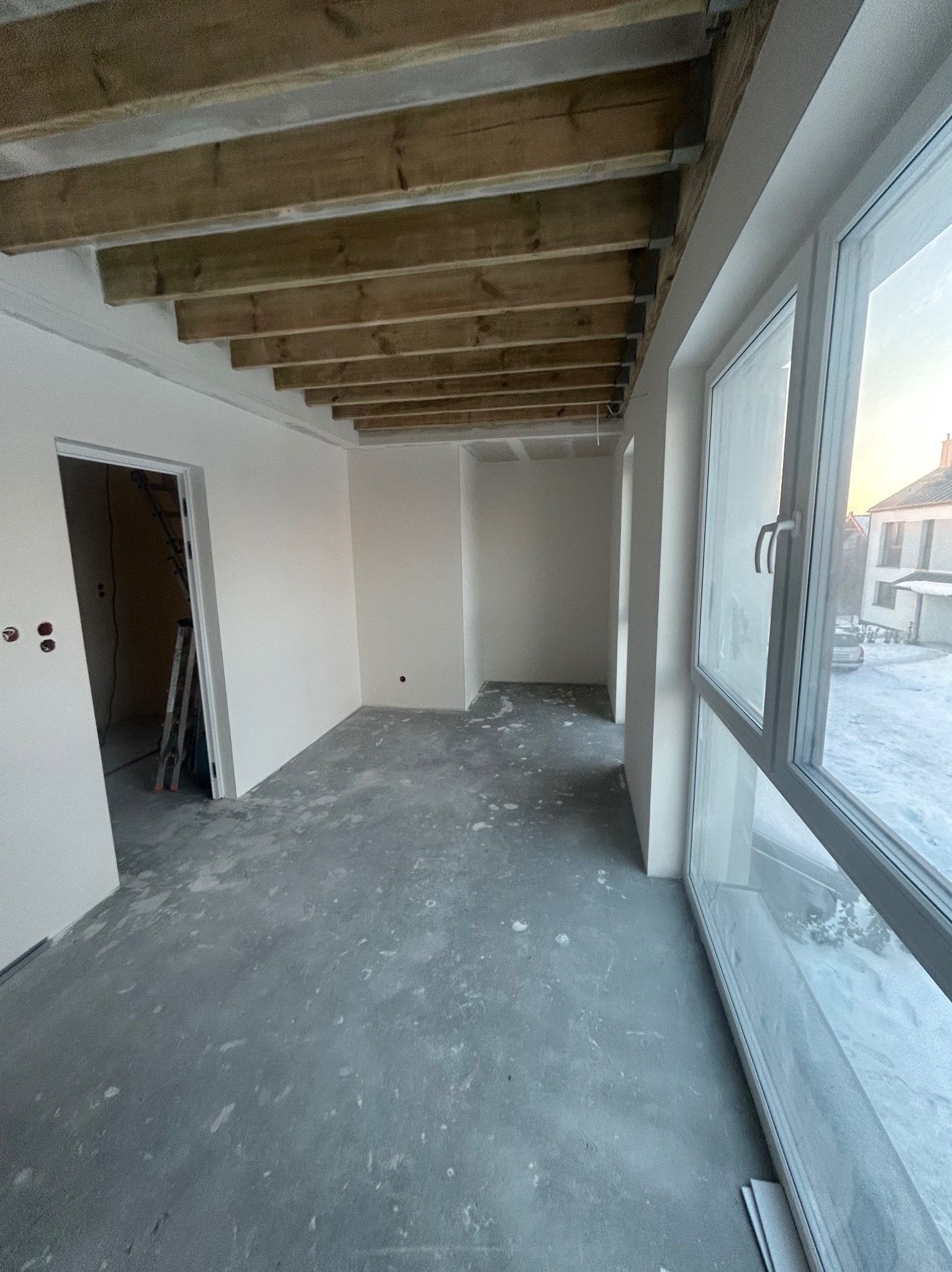 Surowe wnętrze z drewnianym stropem i dużymi oknami. Ściany świeżo pomalowane na biało, podłoga betonowa. Widoczna drabina i instalacja elektryczna.