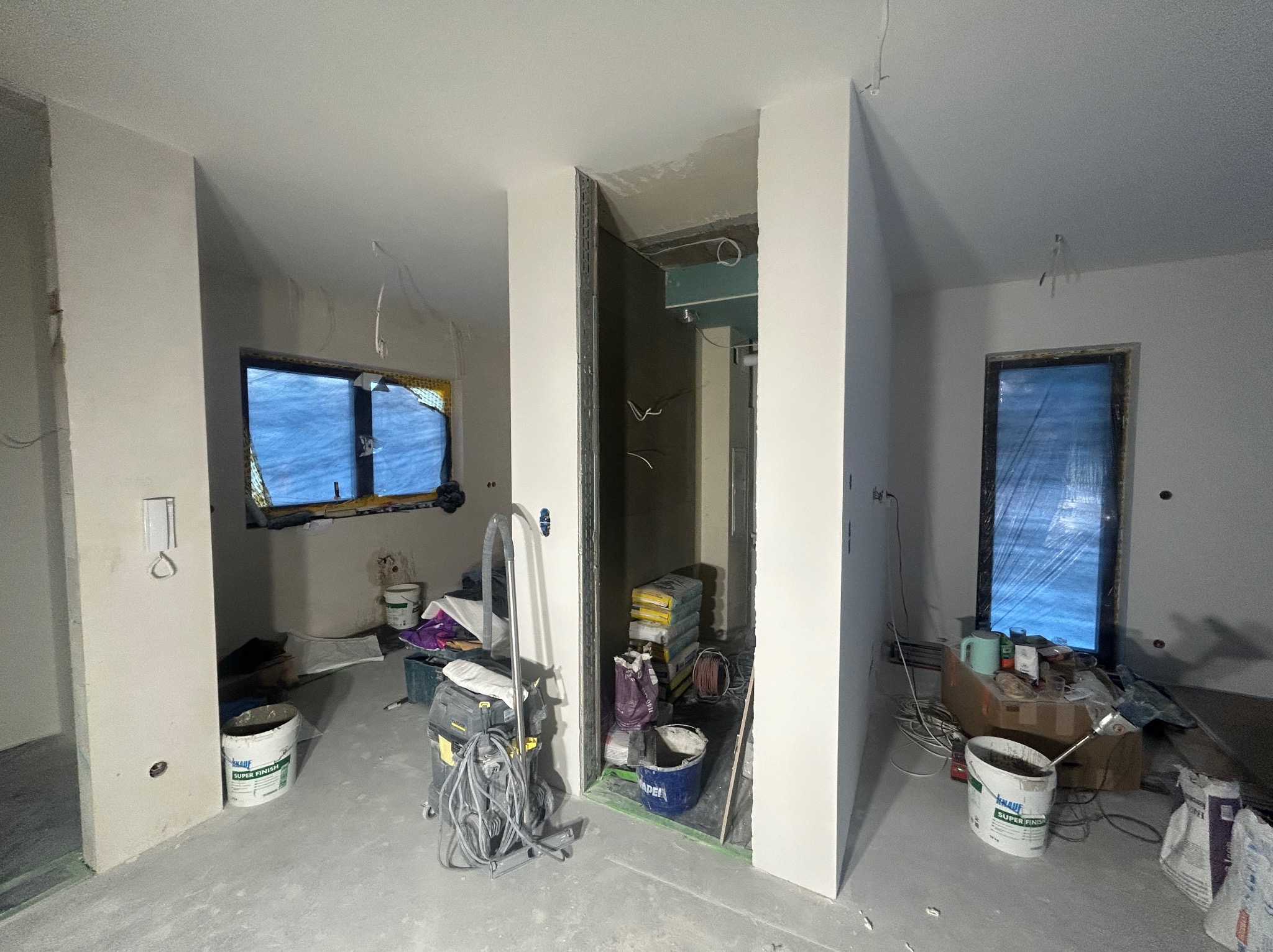 Wnętrze w trakcie prac wykończeniowych: świeżo położone gładzie, okna zabezpieczone folią, widoczne worki z zaprawą i narzędzia. Stan surowy po szpachlowaniu.