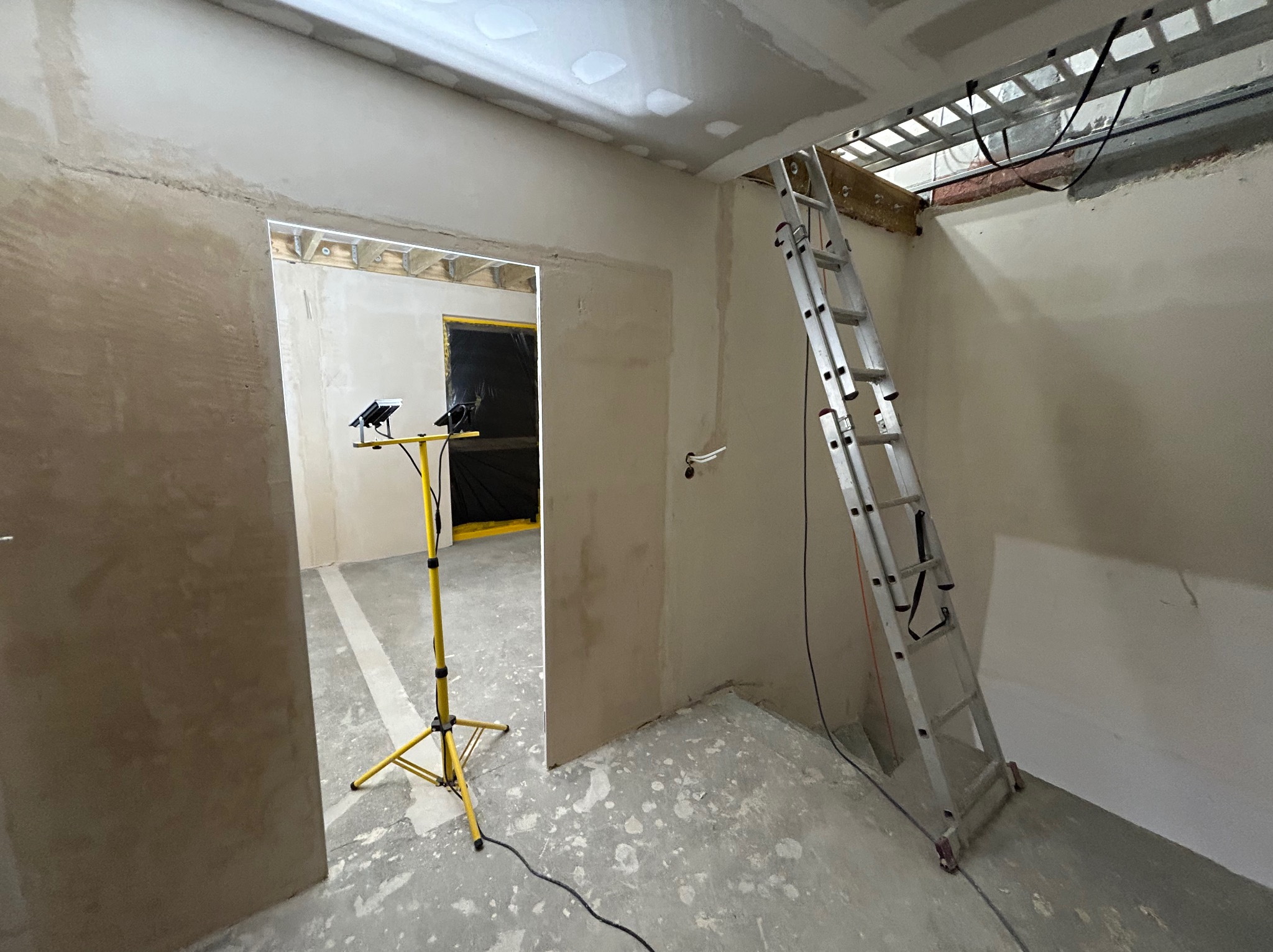 Wnętrze w trakcie prac wykończeniowych: ściany po szpachlowaniu, sufit z płyt karton-gips, drabina oparta o ścianę, lampa budowlana. Widoczne niedoskonałości i detale.
