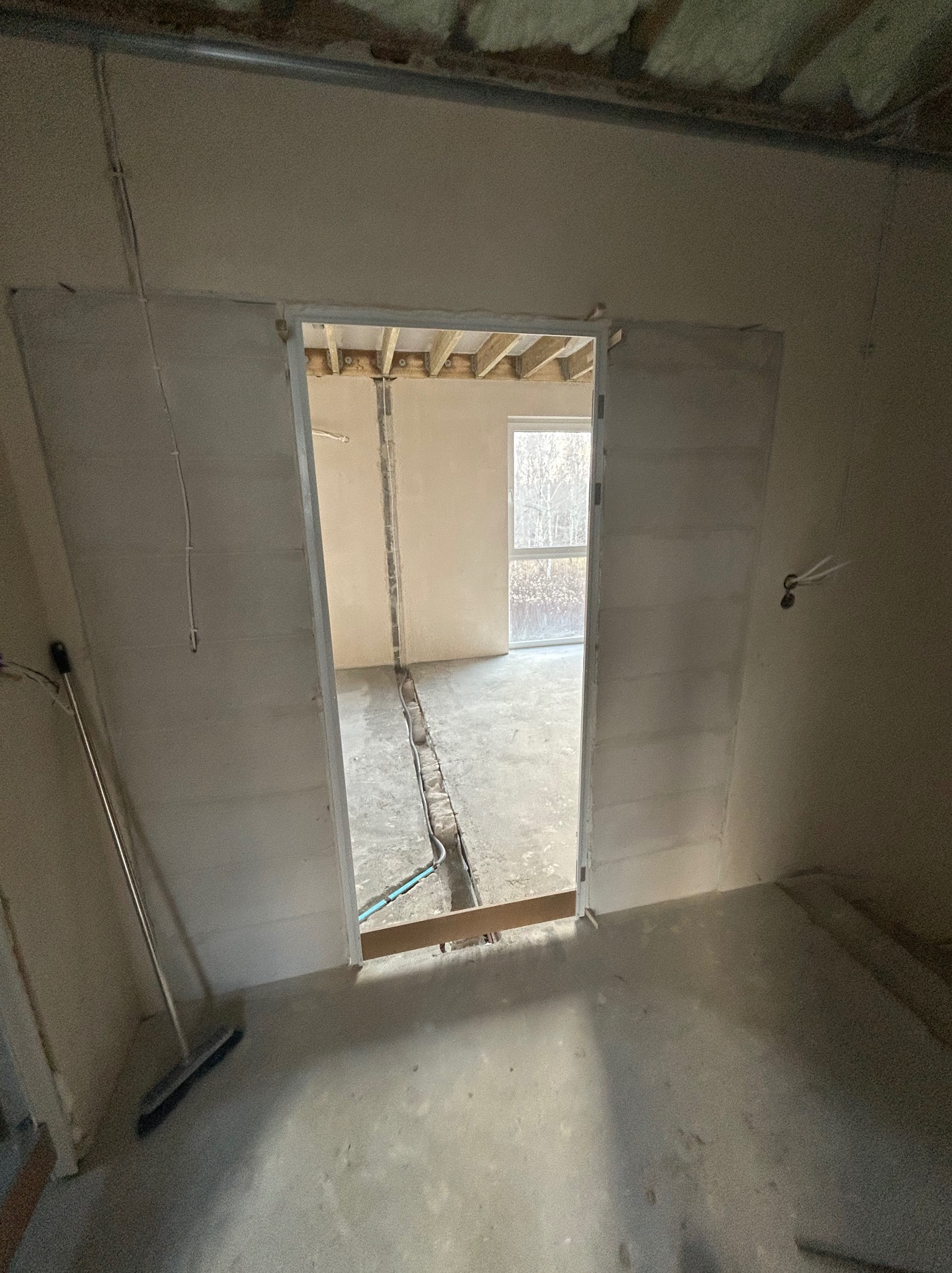 Surowe wnętrze w trakcie prac wykończeniowych: ściany po szpachlowaniu, widoczne instalacje w podłodze, sufit z izolacją. Przejście do kolejnego pomieszczenia z oknem.