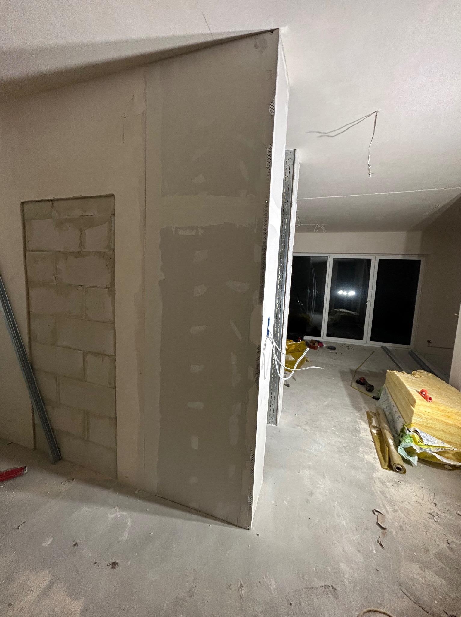 Wnętrze w trakcie prac wykończeniowych: ściany z płyt gipsowo-kartonowych, widoczne profile, instalacja elektryczna, okno balkonowe. Na podłodze materiały izolacyjne.