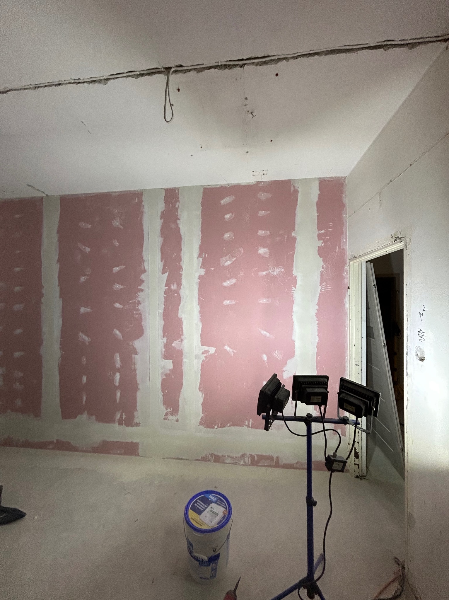 Ściana z płyt karton-gips, częściowo pokryta różowym podkładem i białymi pasami szpachli, oświetlona lampami budowlanymi. Widoczny fragment sufitu i podłogi.