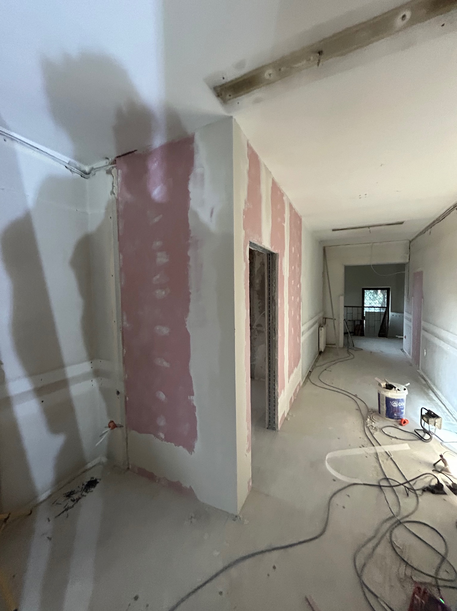 Korytarz w trakcie prac wykończeniowych: ściany z płyt gipsowo-kartonowych, częściowo pokryte różową warstwą, widoczne kable i narzędzia na podłodze, surowy stan.
