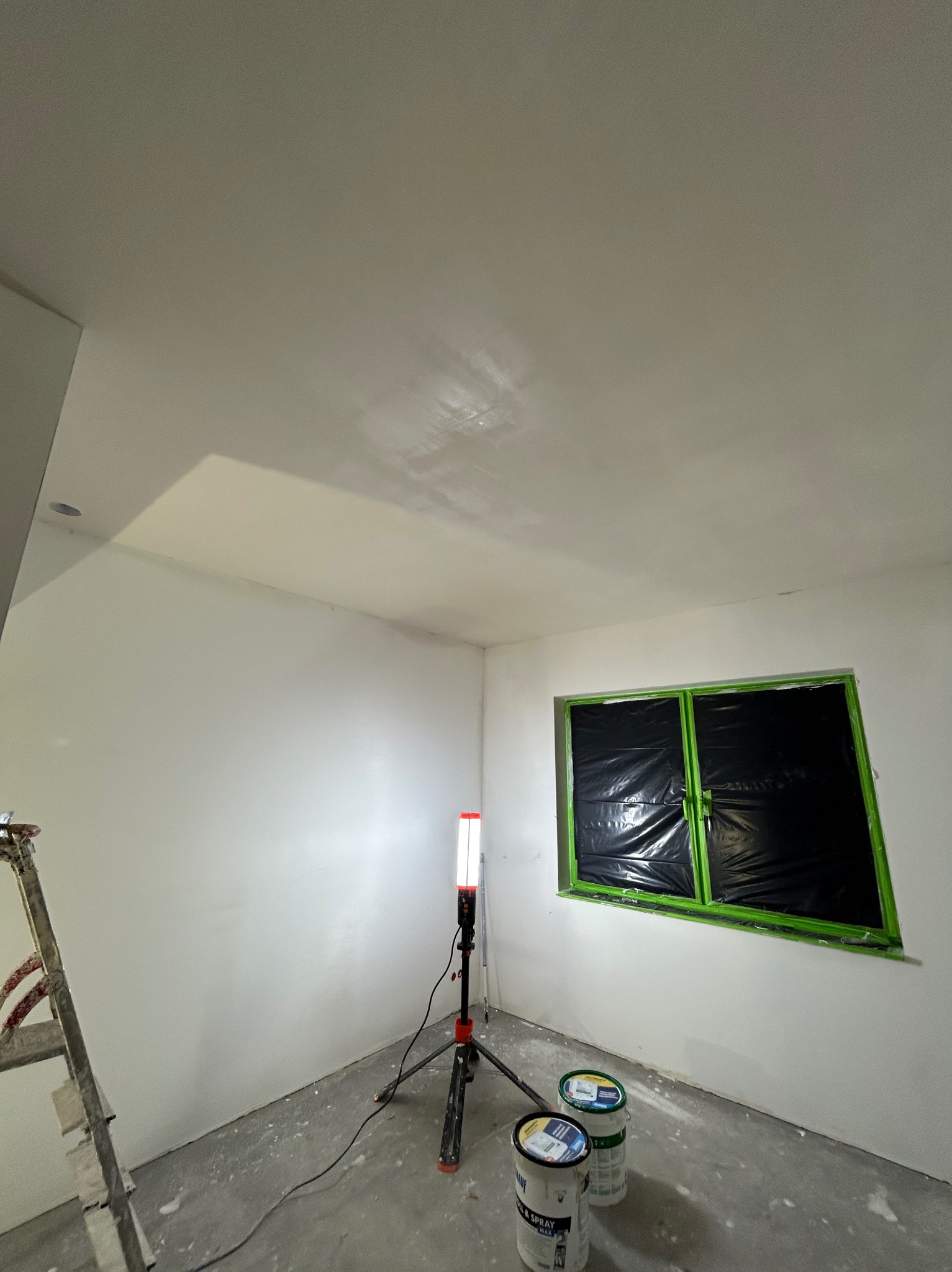 Wnętrze po szpachlowaniu w Łazach: gładkie ściany, zabezpieczone okno folią, drabina i lampa budowlana. Przygotowanie do malowania.